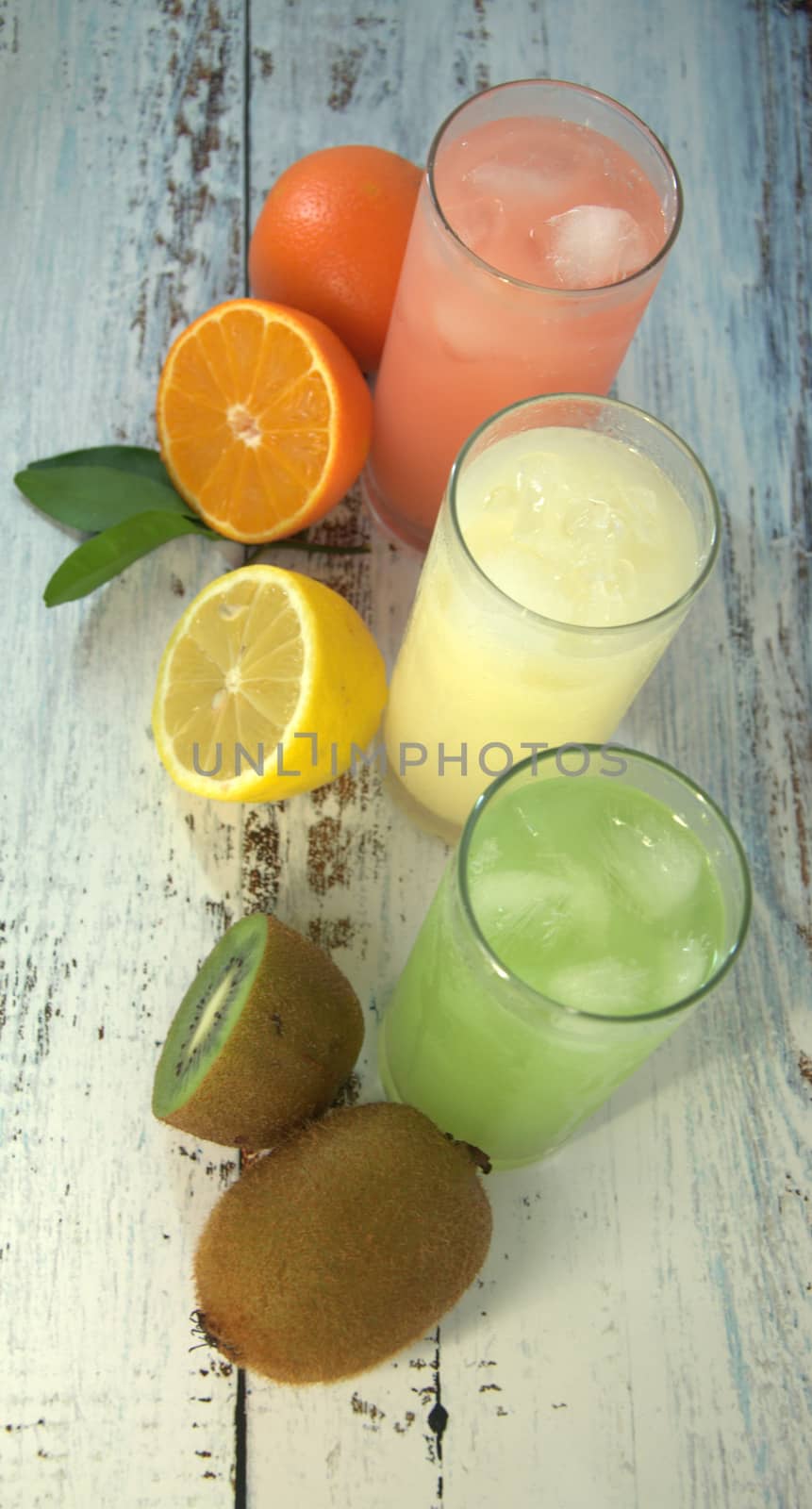 Three glass glasses with a refreshing juice and ice, on a textile stand, whole and sliced half of an orange with leaves, lemon and kiwi, lay on a white wooden table. Close-up.