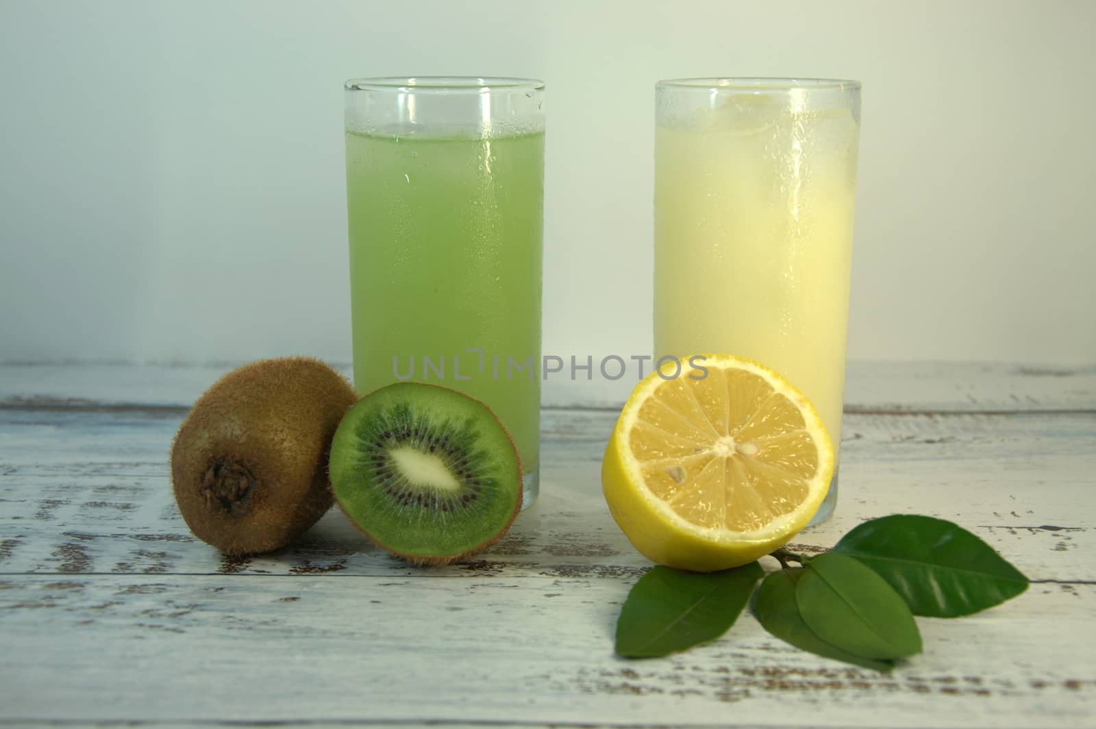 Two glass glasses with a refreshing juice and ice, on a textile stand, whole and sliced half of an orange with leaves and kiwi, lies on a white wooden table. by alexey_zheltukhin