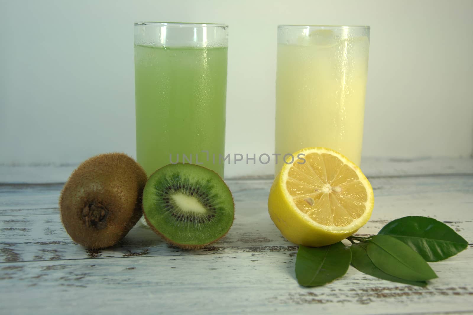 Two glass glasses with a refreshing juice and ice, on a textile stand, whole and sliced half of an orange with leaves and kiwi, lies on a white wooden table. Close-up.