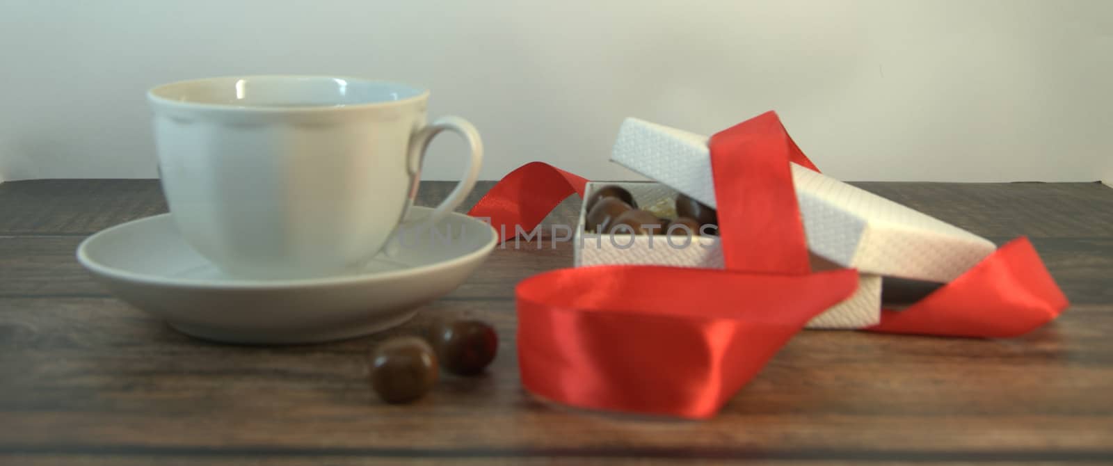 A cup of tea, a white box of chocolates with a red satin ribbon on a wooden table. Close-up.