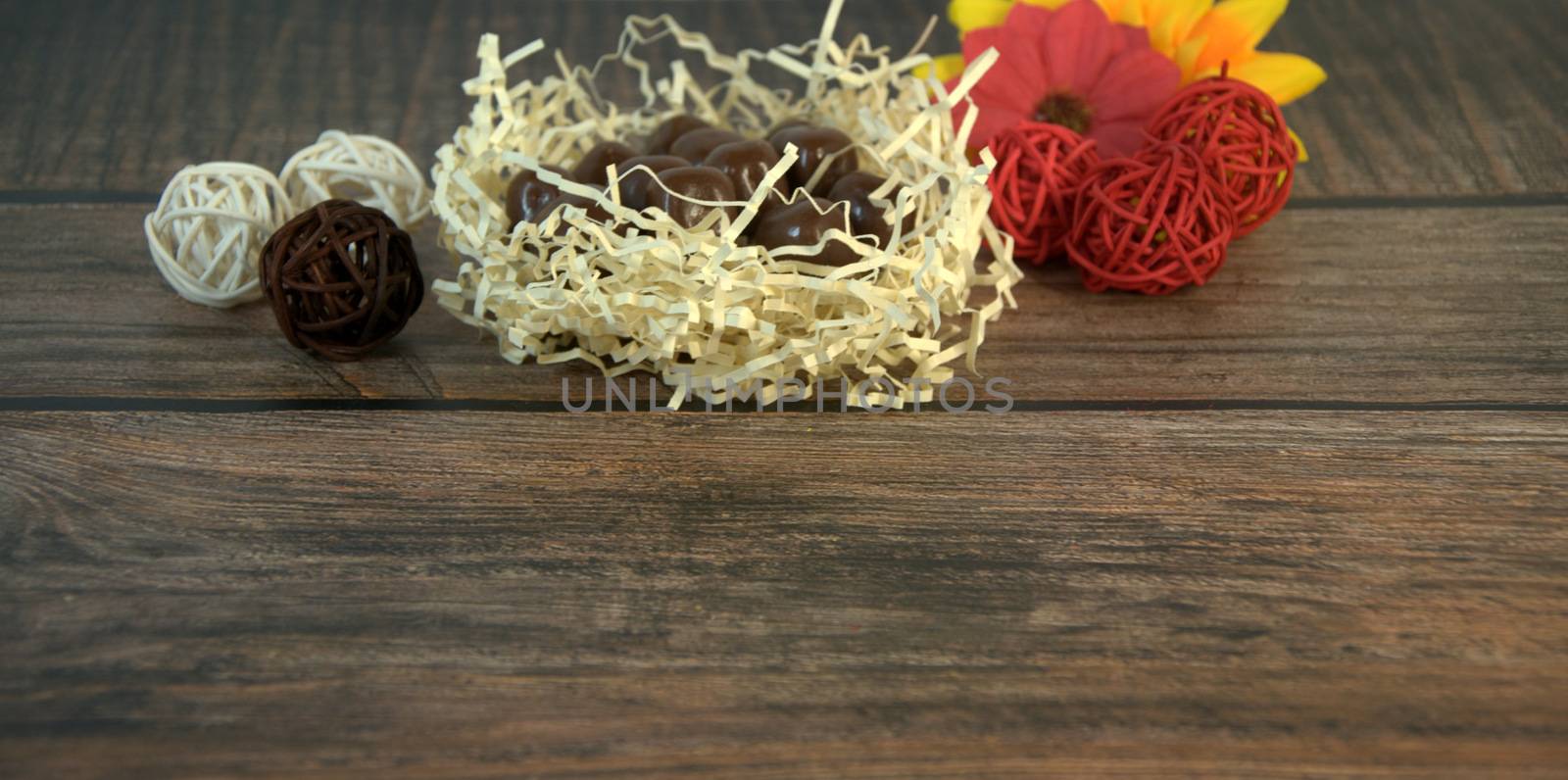 Round chocolates in a nest of straw, multicolored decorative balls and flower buds on a wooden table. Close-up.