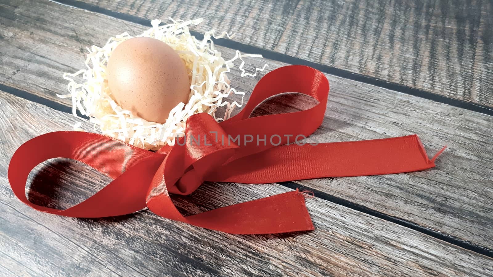 Chicken egg in a nest of straw and red satin ribbon on a wooden table. by alexey_zheltukhin