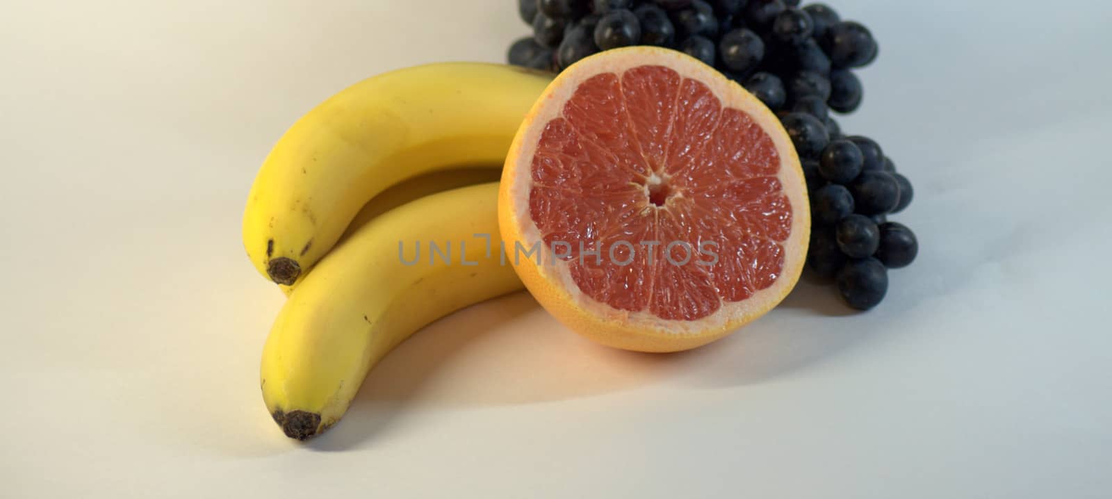 A bunch of ripe, tasty bananas, three pieces, a bunch of grapes and half a ripe grapefruit. The picture was taken in the lightbox, close-up.
