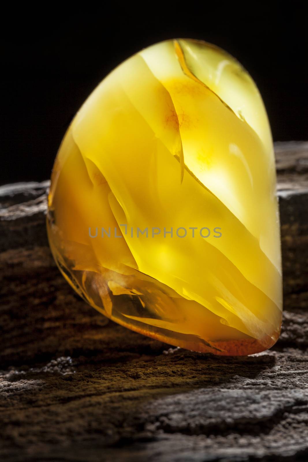 A piece of yellow semi opaque natural amber, classification color Bastard, has cracks inside. Polished, has a bead shape. Placed on dark stoned wood texture.