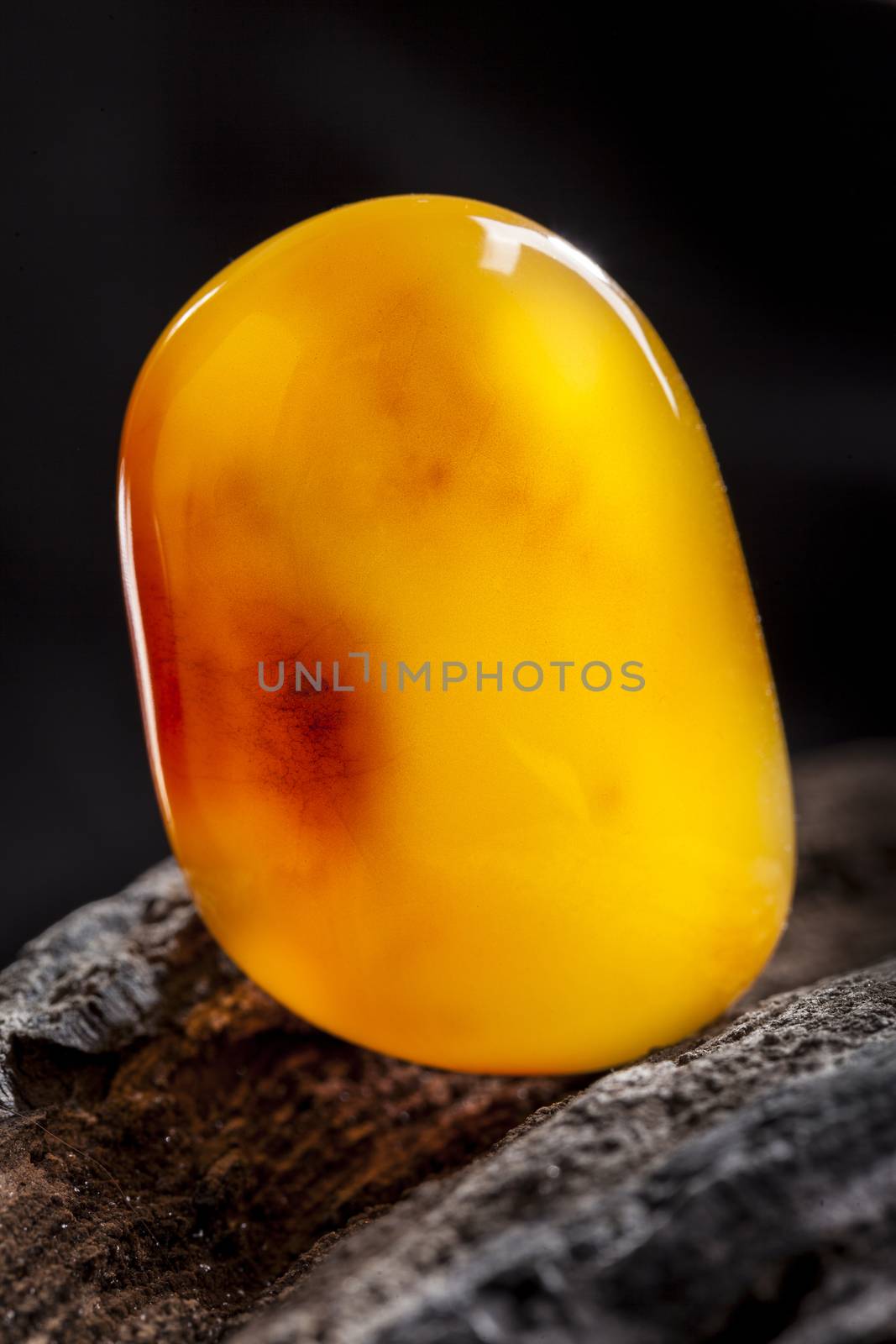 A piece of yellow semi opaque natural amber, classification color Bastard, has cracks inside. Polished, has a bead shape. Placed on dark stoned wood texture.