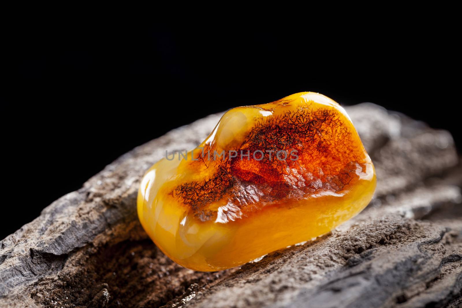 A piece of yellow semi opaque natural amber, classification color Bastard, has cracks inside. Polished, has a bead shape. Placed on dark stoned wood texture.