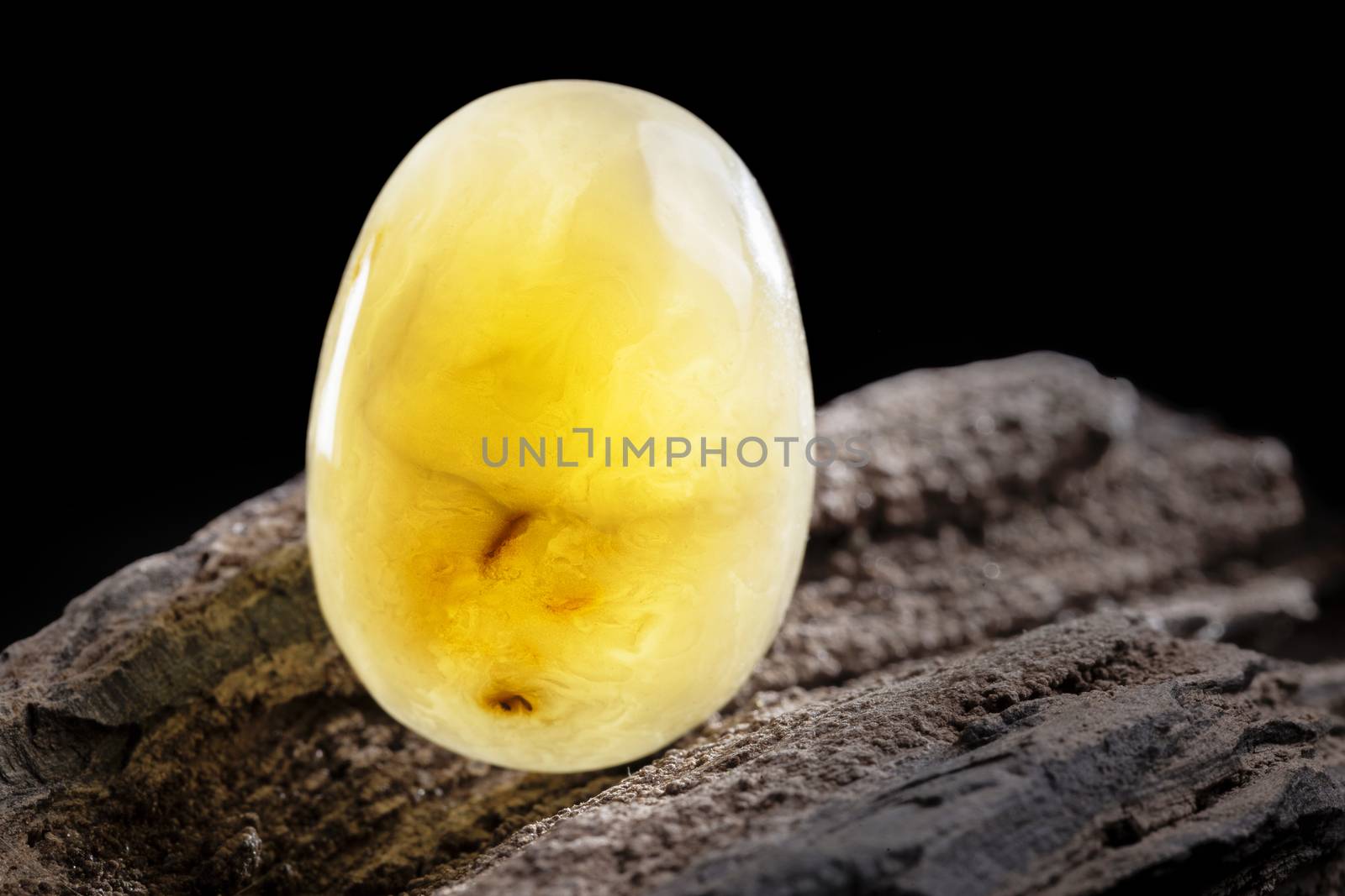 A piece of yellow semi opaque natural amber, classification color Bastard, has cracks inside. Polished, has a bead shape. Placed on dark stoned wood texture.