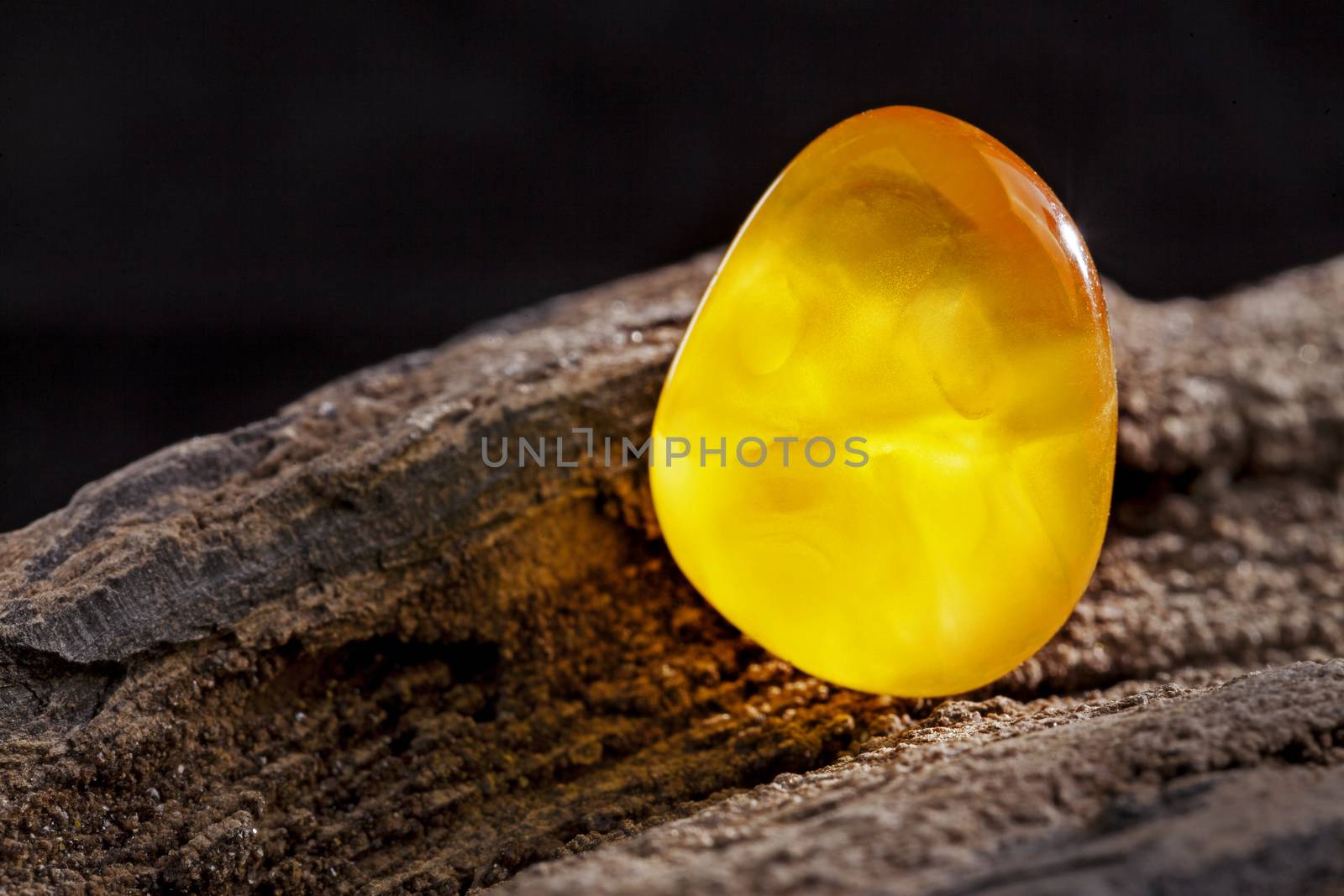 A piece of yellow semi opaque natural amber, classification color Bastard, has cracks inside. Polished, has a bead shape. Placed on dark stoned wood texture.