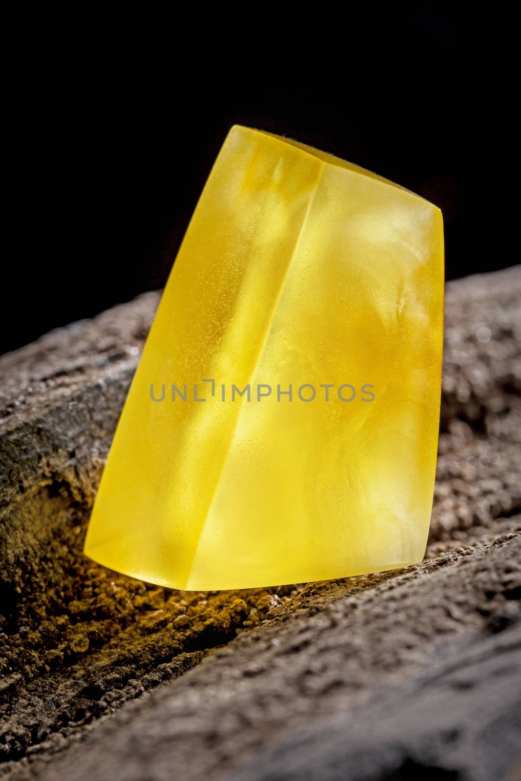 A piece of yellow semi opaque natural amber, classification color Bastard, has cracks inside. Polished, has a bead shape. Placed on dark stoned wood texture.