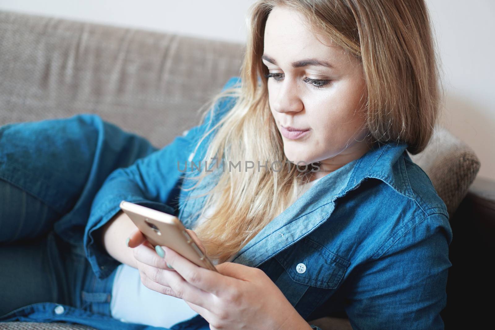 Portrait of thoughtful woman lying on couch pillow holding smart phone in hands looking at screen