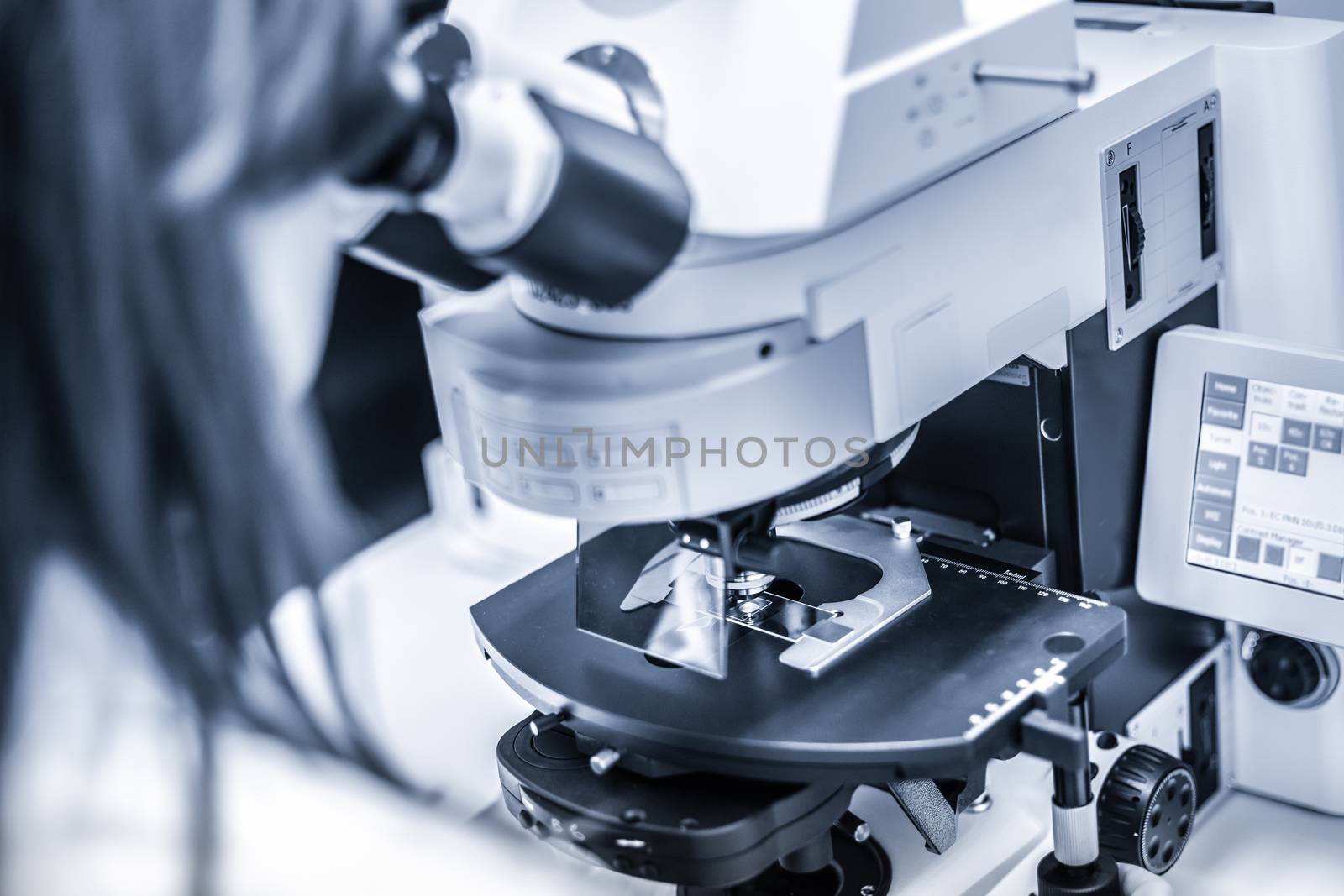 Female scientist microscoping on hi-tec fluorescent microscope. Health care professional in hes working environment. Blue toned grayscale image.