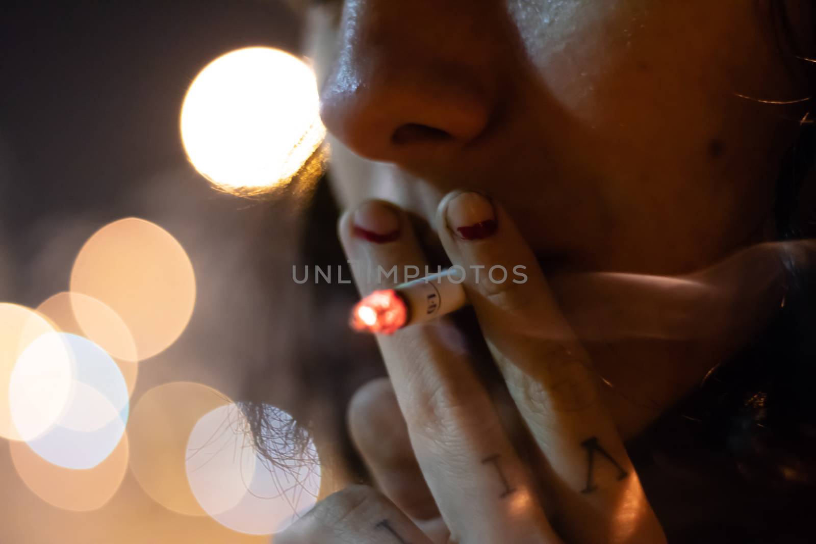 a girl smoking a cigarette with rubbed out red nail polish. background is blurry.