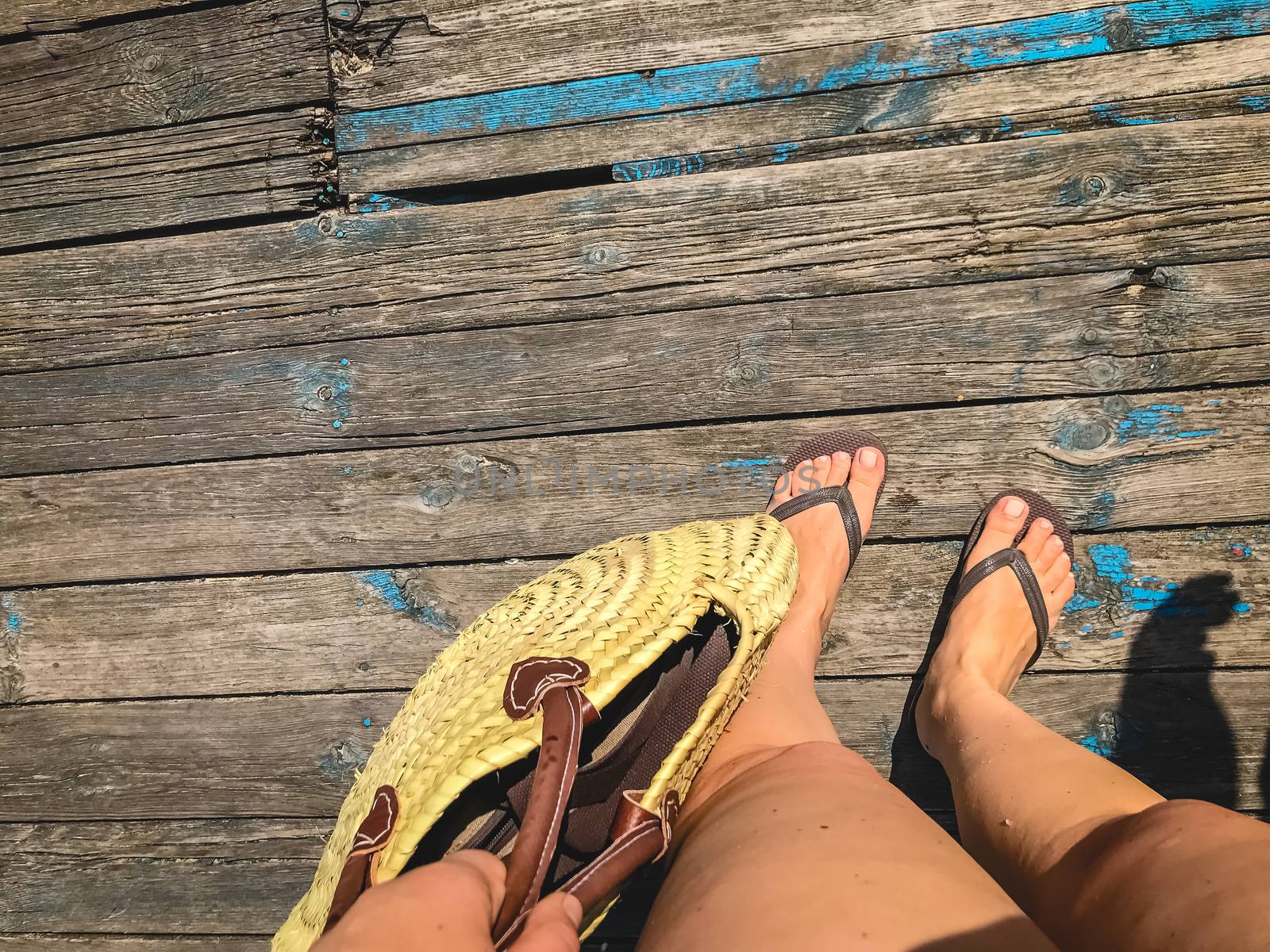 Top view, photo of legs in beach flip-flops and with a straw bag in hand on a wooden old floor. Photos on vacation, beach, summer.