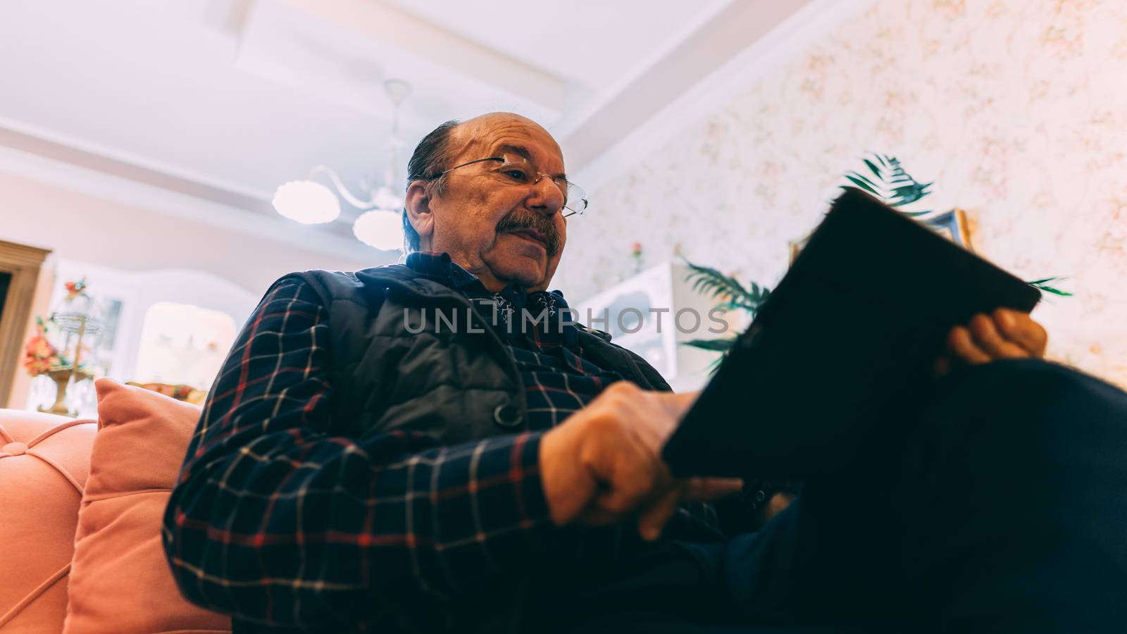 Senior man with tablet sitting on sofa at home. by Emurado