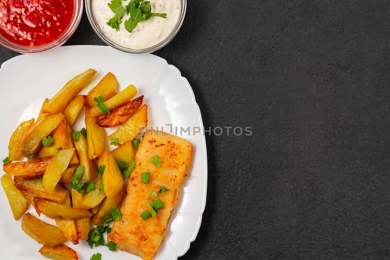 Fried fish and chips on a white plate on the black kitchen table with tomato sauce, tartar sauce and pickled vegetables salad - photo, image. Flat lay, top view with copyspace by galsand
