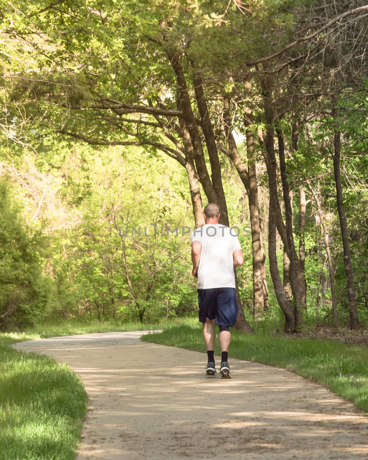 Rear view of senior Caucasian man running at nature park during sunset. Healthy person in white crew shirt and long sock running on concrete pathway in natural area near Dallas, Texas, USA