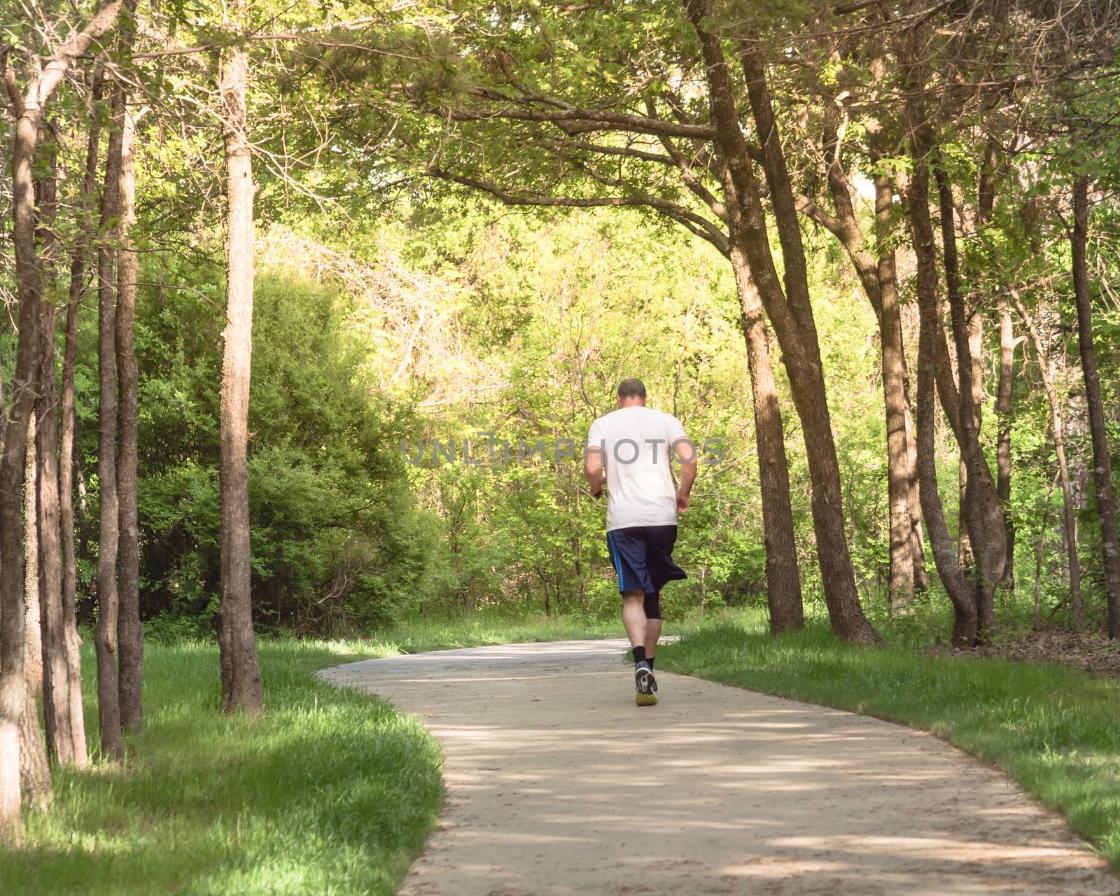 Rear view of senior Caucasian man running at nature park during sunset. Healthy person in white crew shirt and long sock running on concrete pathway in natural area near Dallas, Texas, USA