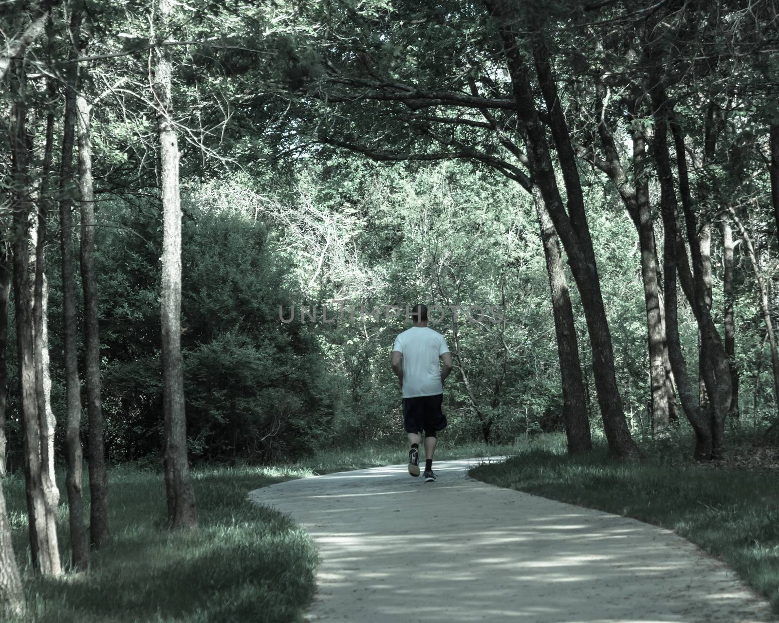 Senior Caucasian man running at nature park during sunset. Rear view of healthy person in white crew shirt and long sock running on s-curved concrete pathway in natural area near Dallas, Texas, USA