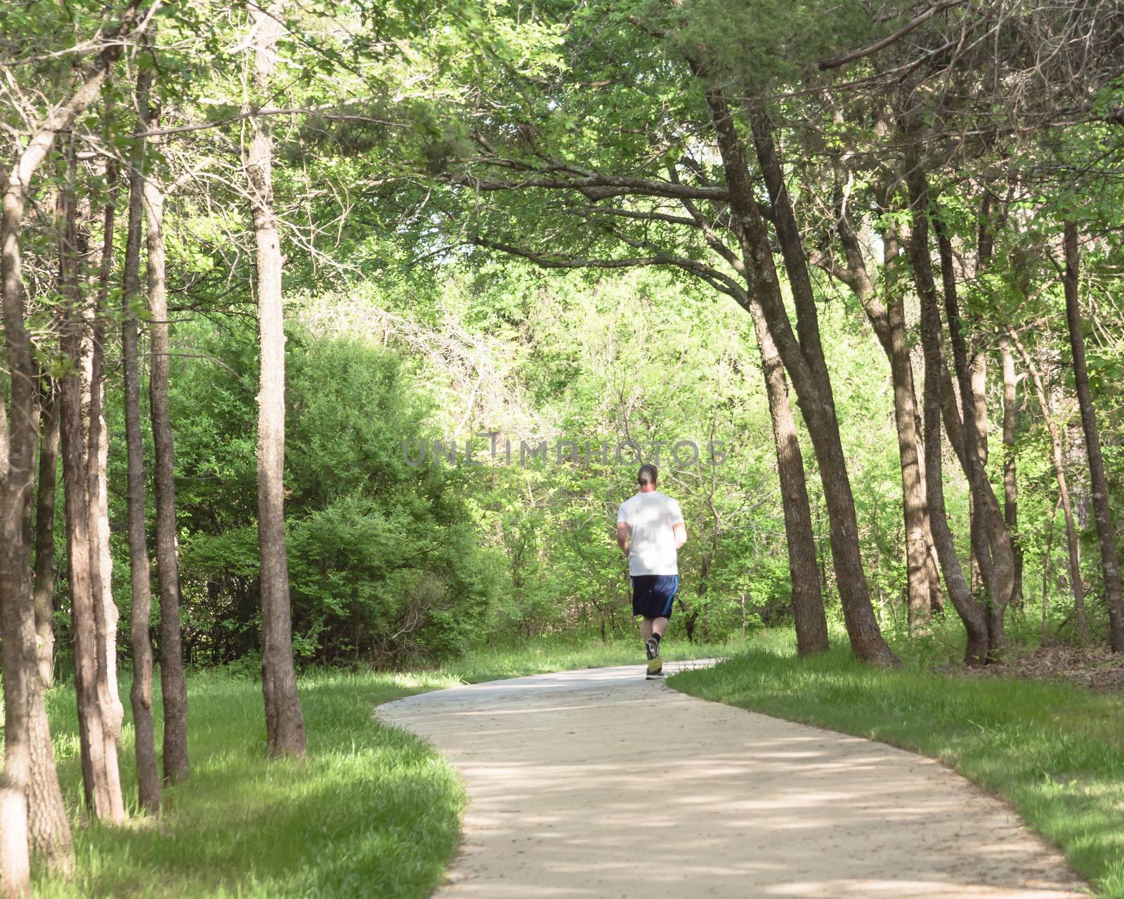 Senior Caucasian man running at nature park during sunset. Rear view of healthy person in white crew shirt and long sock running on s-curved concrete pathway in natural area near Dallas, Texas, USA