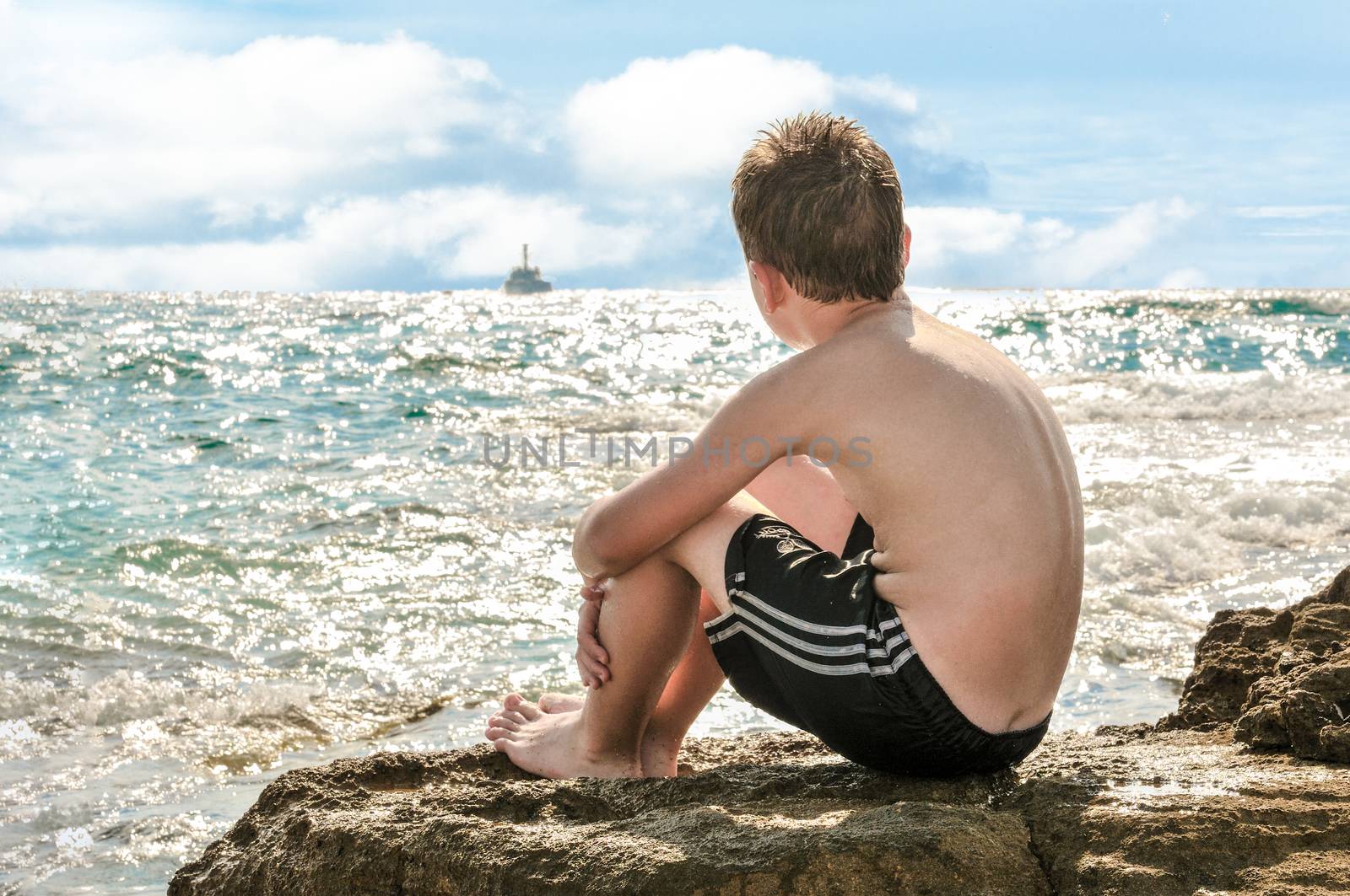 
	
The teenager sits on a large rock and looks at the ship leaving the horizon.