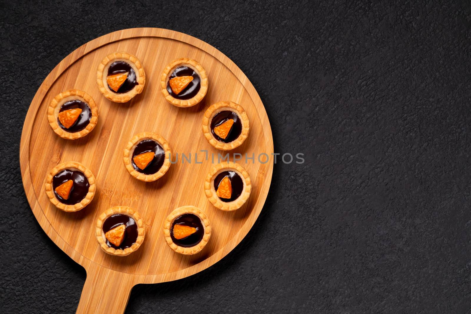 Sweet tartlets with chocolate and slices of tangerine on a wooden dish for serving, top view.