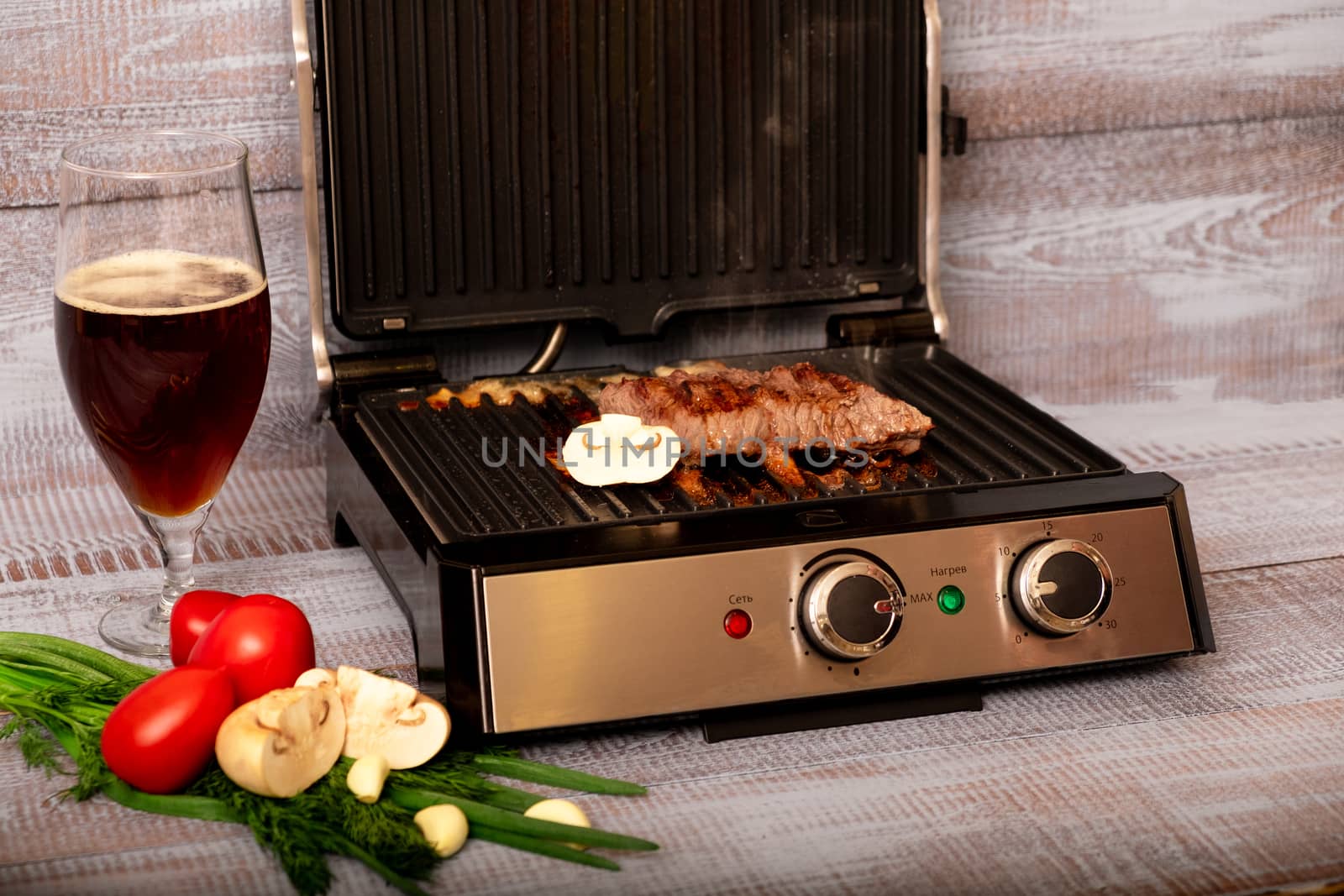 Beef is cooked on the electric grill. Around the grill vegetables laid out. On a wooden background.