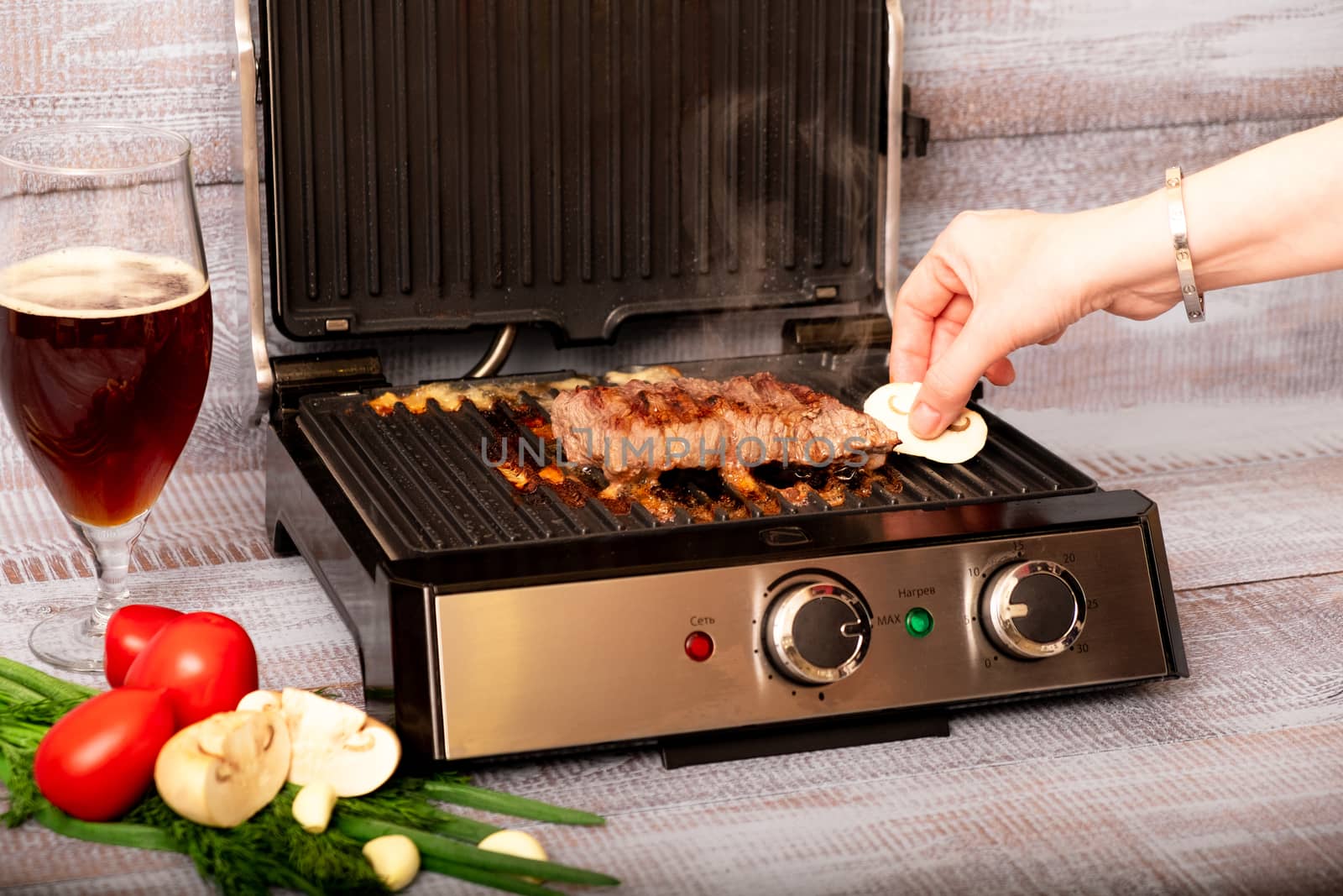 Beef is cooked on the electric grill. Around the grill vegetables laid out. On a wooden background.