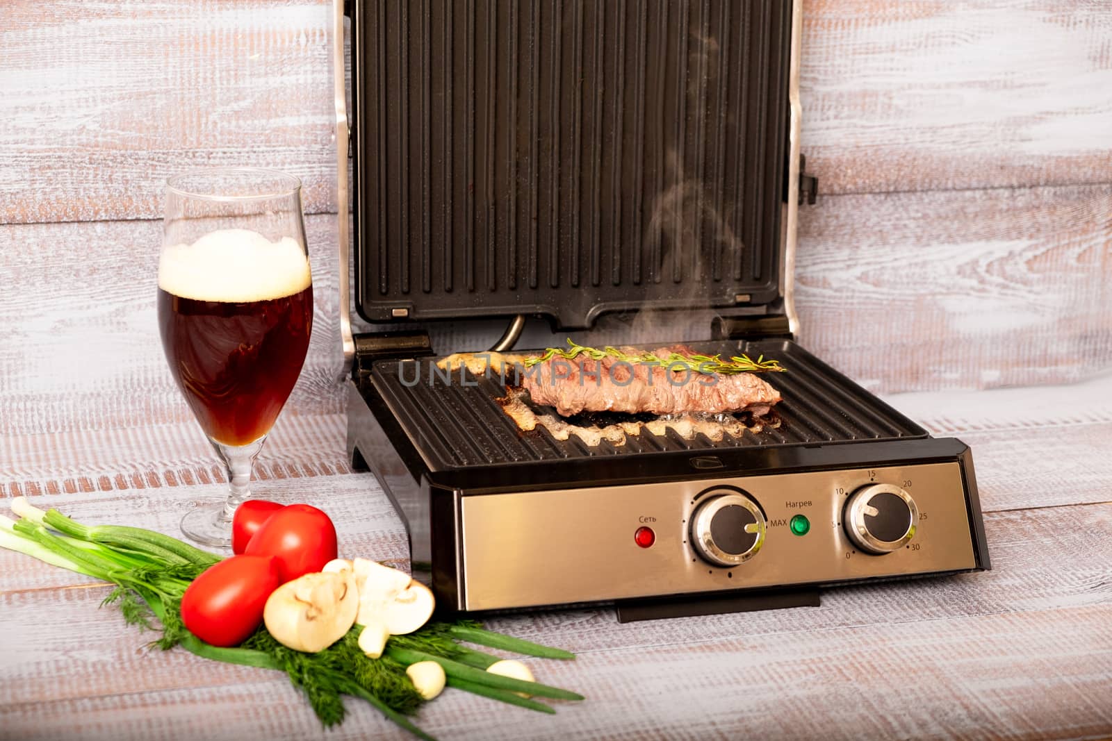 Beef is cooked on the electric grill. Around the grill vegetables laid out. On a wooden background.