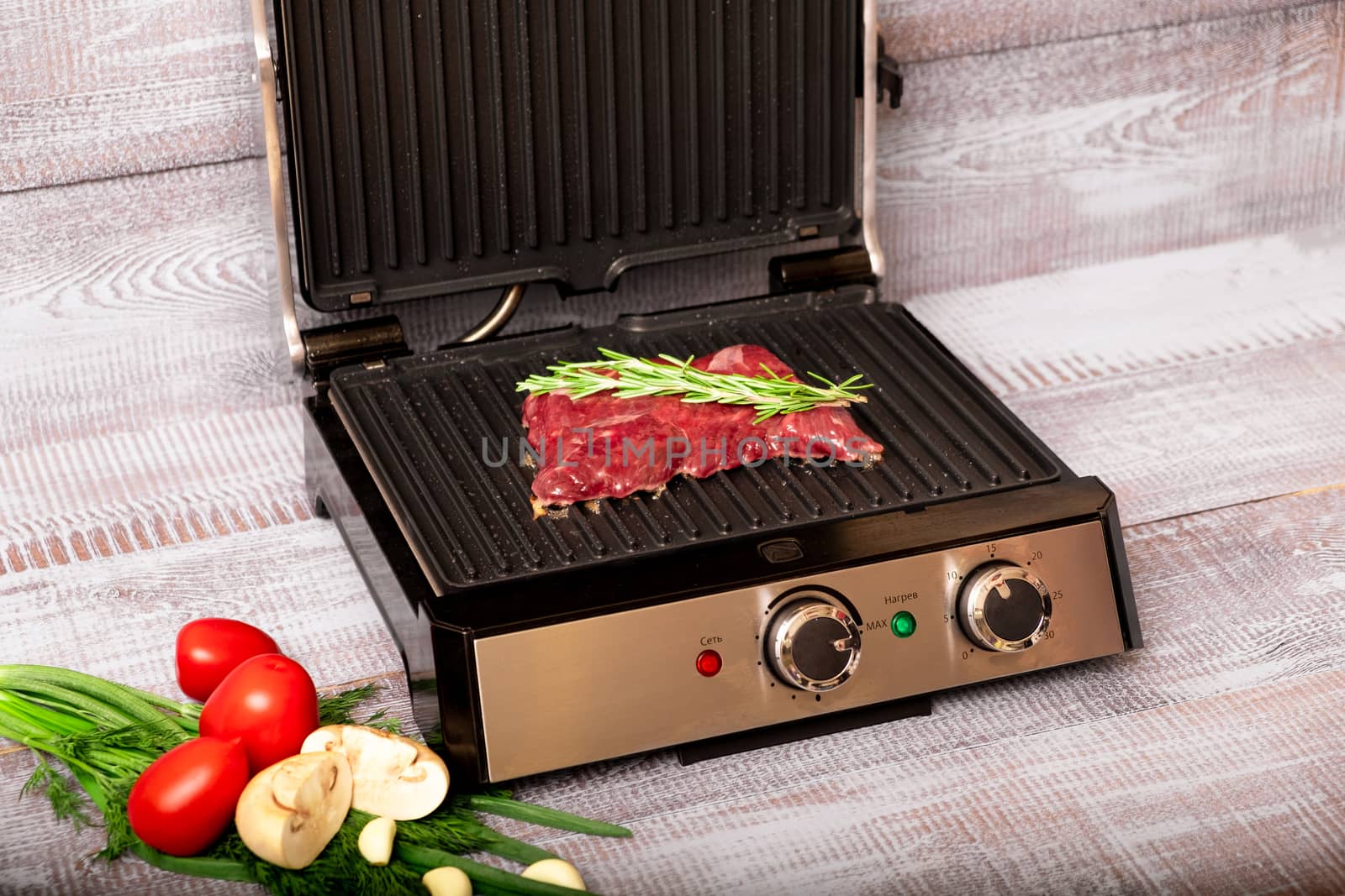 Beef is cooked on the electric grill. Around the grill vegetables laid out. On a wooden background.