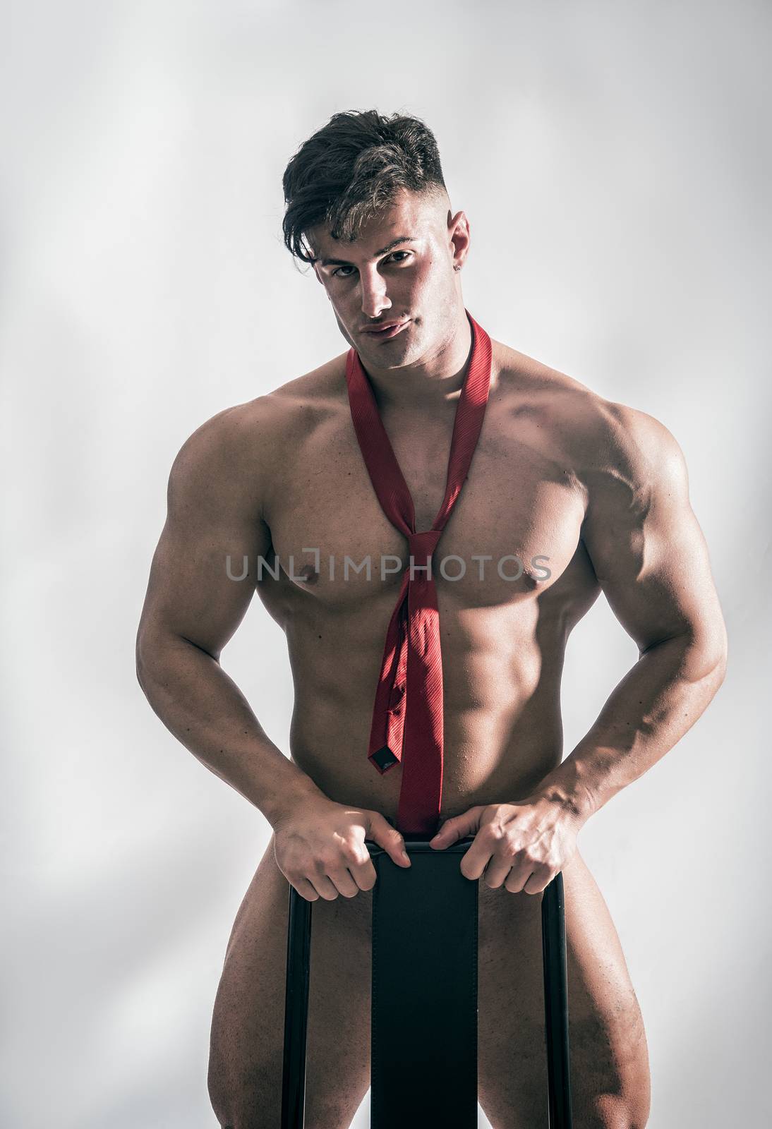 Muscular bodybuilder standing behind chair, totally naked, looking at camera