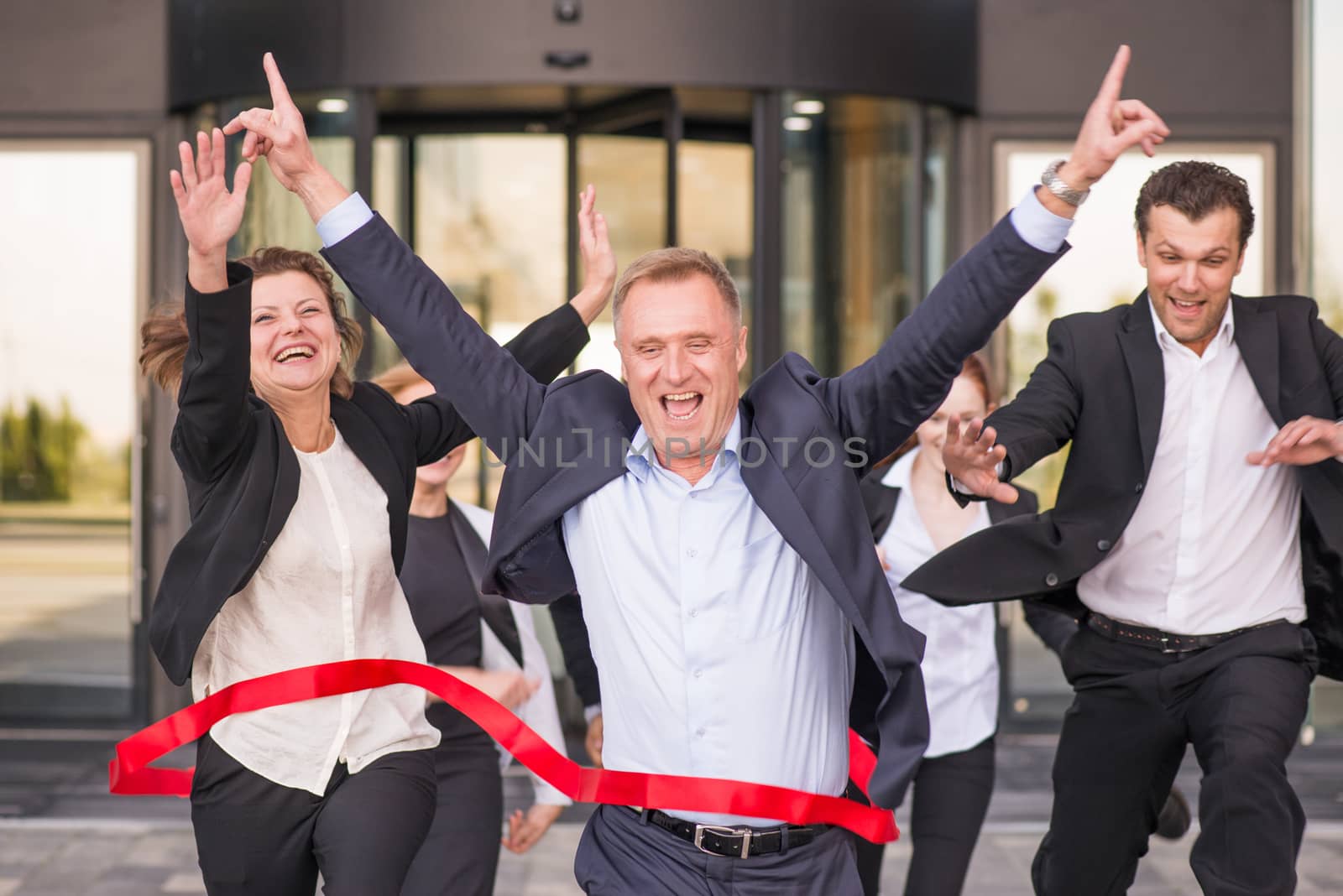 Group of happy business people running from office building crossing red ribbon finish line