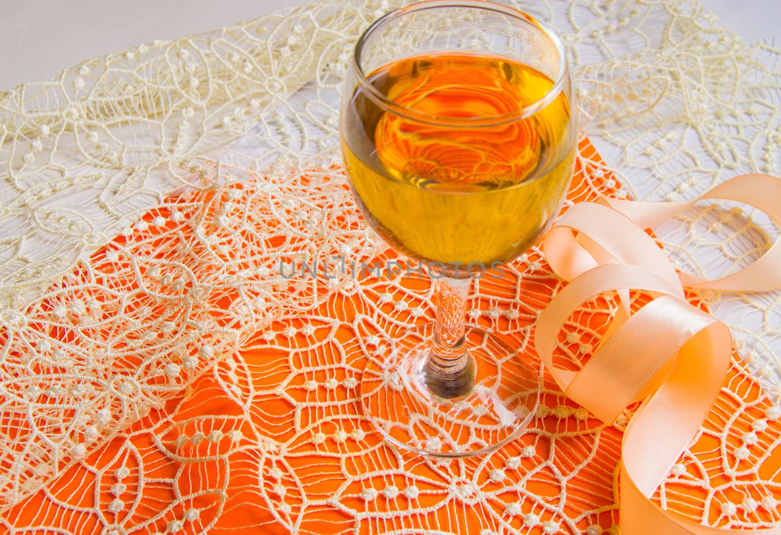 Flat stacking of white wine in a glass, beige lace and decorative ribbon, orange background, top view. Romantic spring or summer horizontal composition. Selective focus.