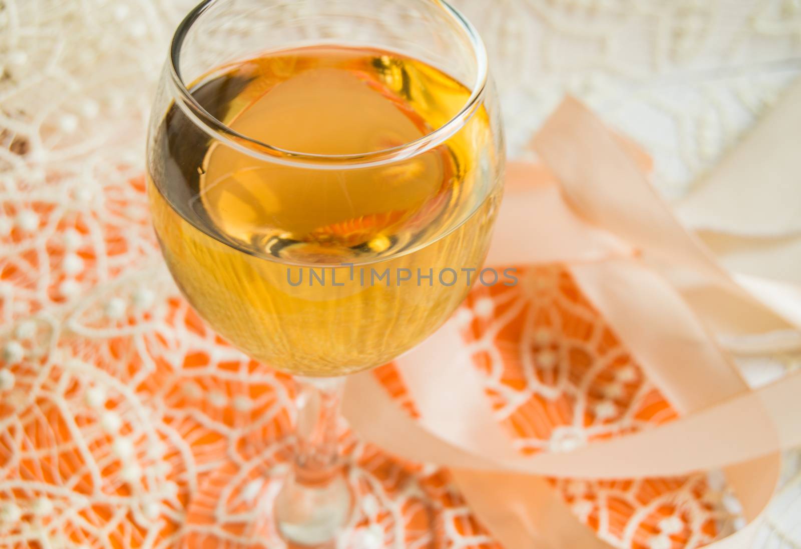 Flat stacking of white wine in a glass, beige lace and decorative ribbon, orange background, top view. Romantic spring or summer horizontal composition. Selective focus.