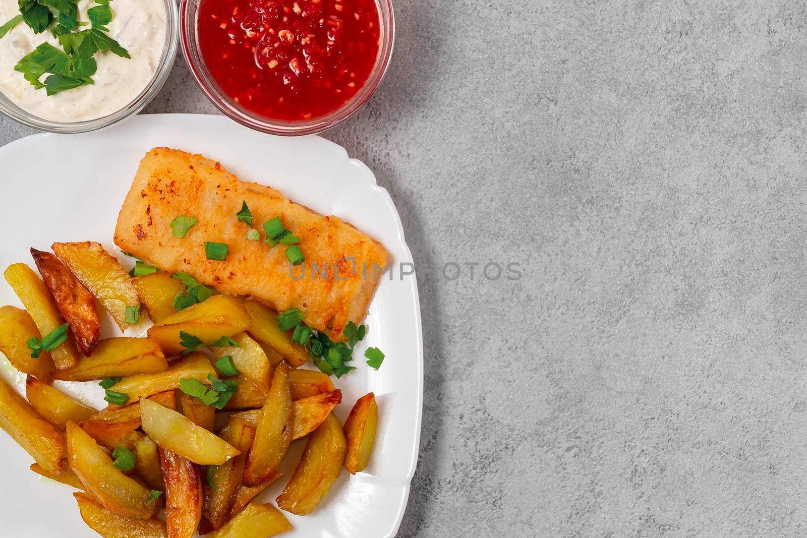 Fried fish and chips on a white plate on the gray kitchen table with tomato sauce, tartar sauce and pickled vegetables salad - photo, image. Flat lay, top view with copyspace by galsand