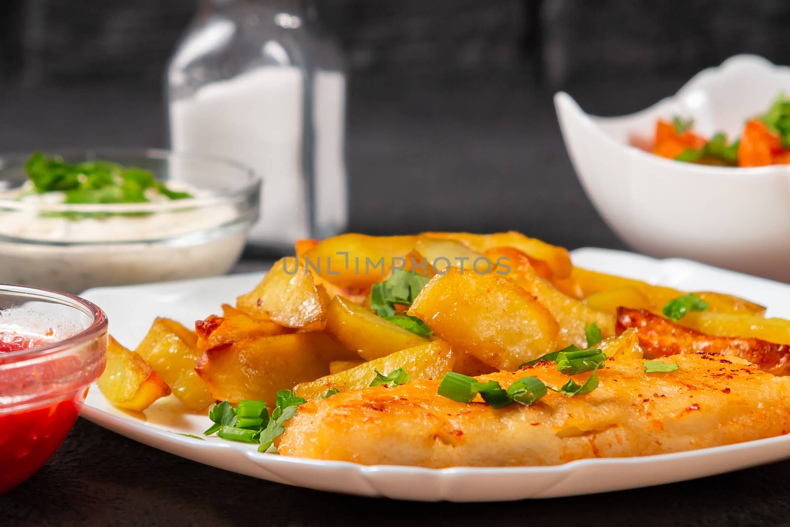 Fried fish and chips on a white plate on the black kitchen table with tomato sauce, tartar sauce and pickled vegetables salad - photo, image by galsand