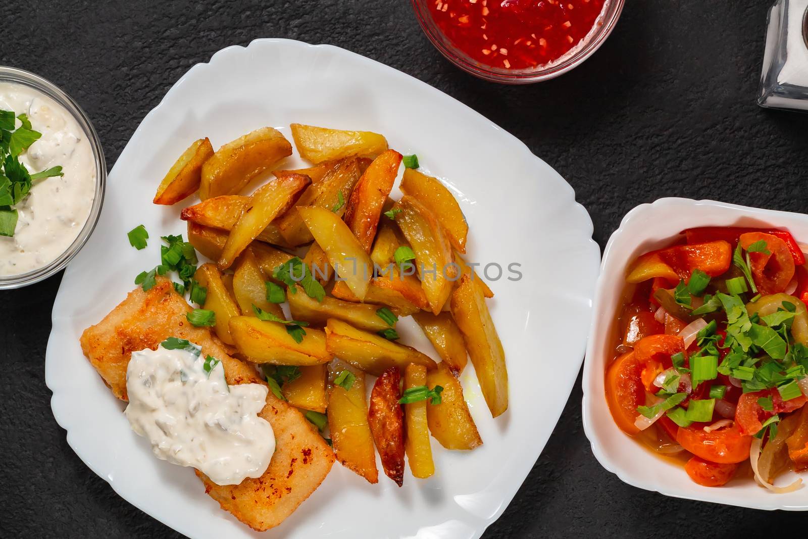 Fried fish and chips on a white plate on the black kitchen table with tomato sauce, tartar sauce and pickled vegetables salad - photo, image. Flat lay, top view