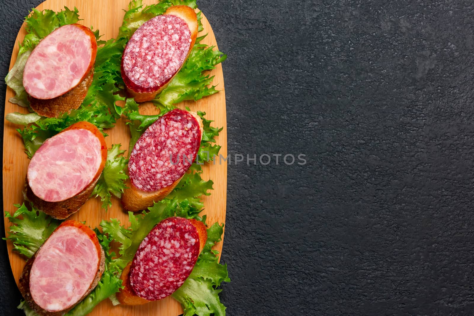 Several sandwiches with sausage and salami and sauce on a black board, flat lay background with copyspace.