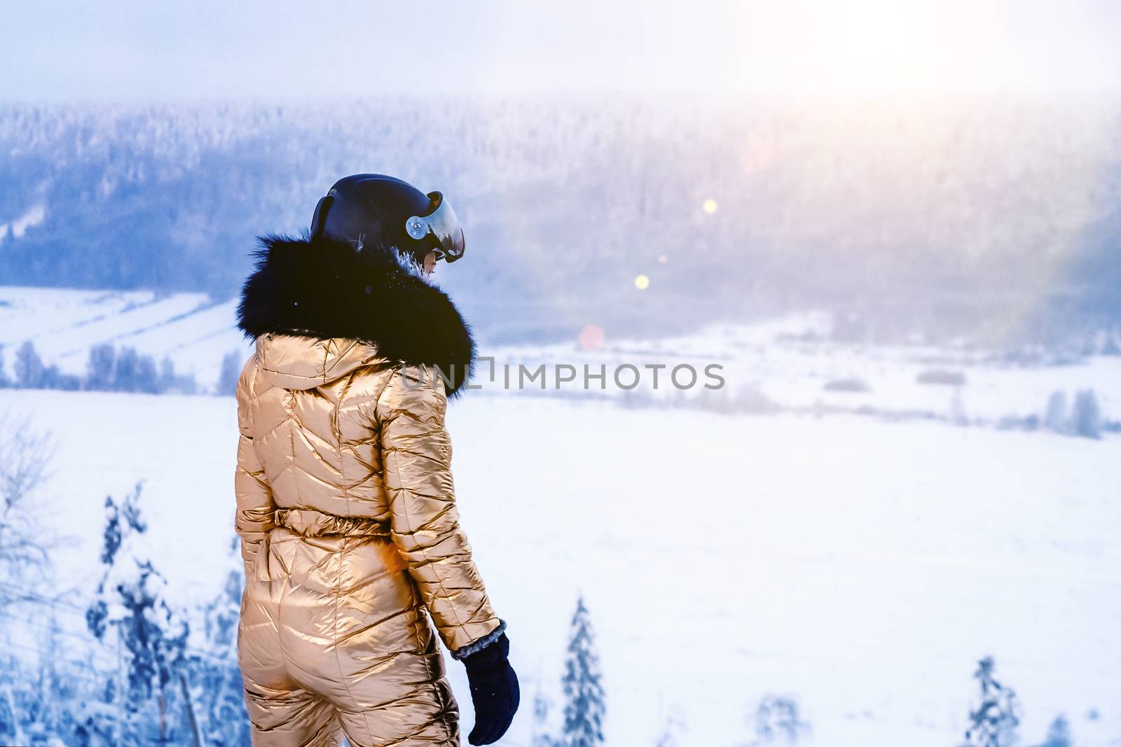 young woman in a golden ski overalls and helmet stands on top of a slope and looks into the distance by galsand