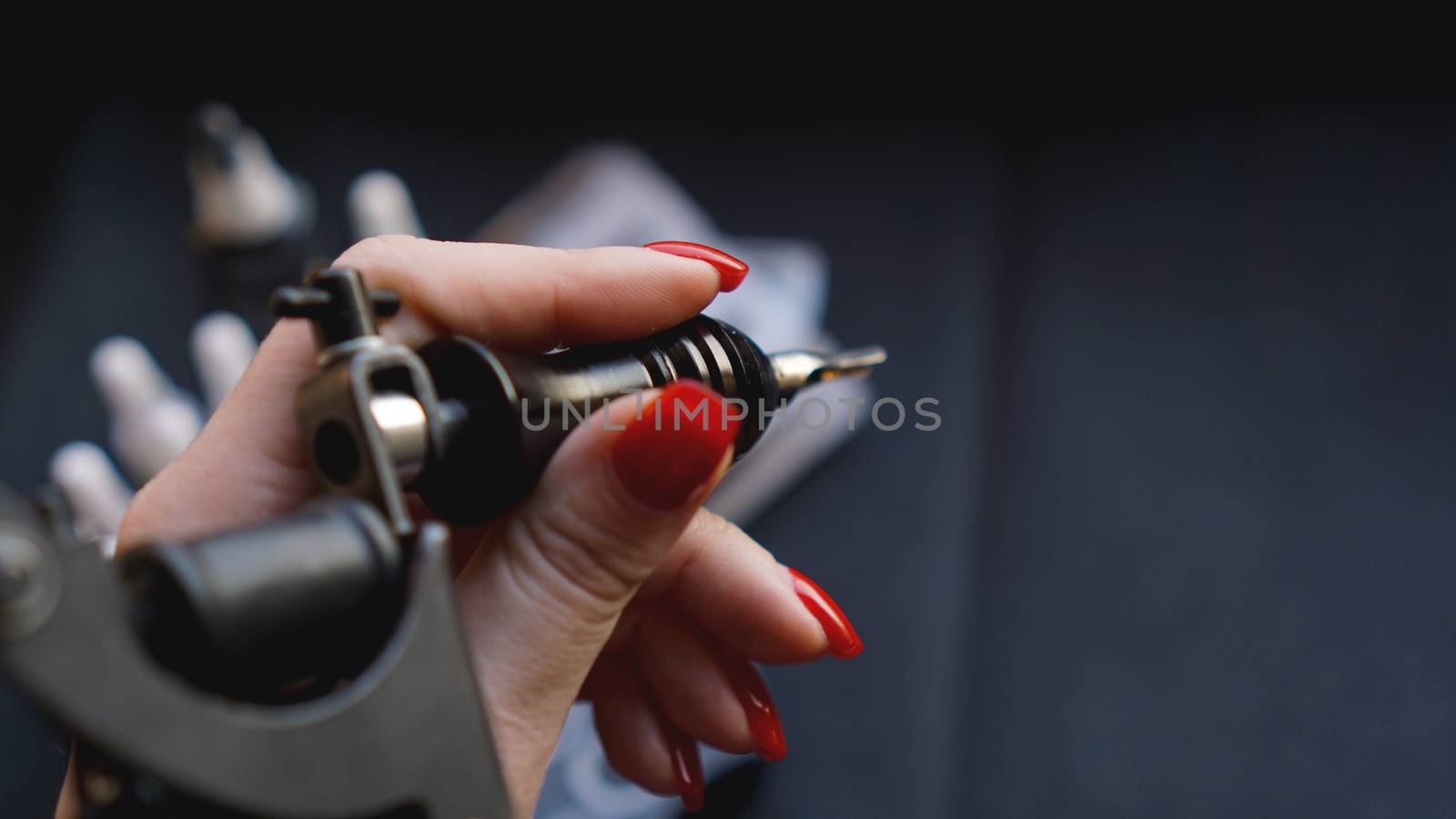 Female hand with red nails holding tattoo machine. Hand without gloves - practice and training tattoo. Dark background