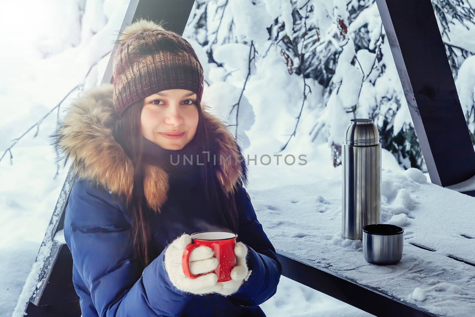 Young girl with a cup of hot coffee in her hands on a bench in the winter snow-covered forest by galsand