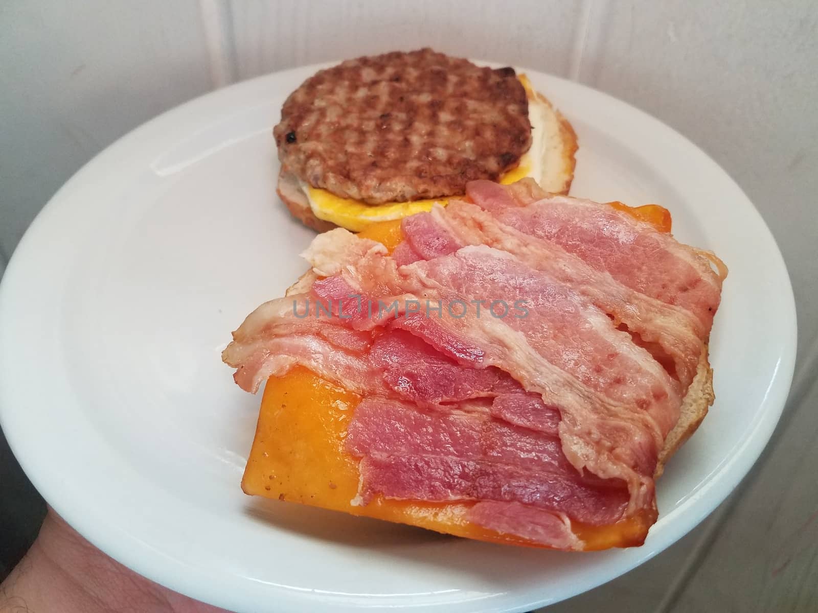 hand holding sausage and egg and bacon and cheese sandwich on white plate