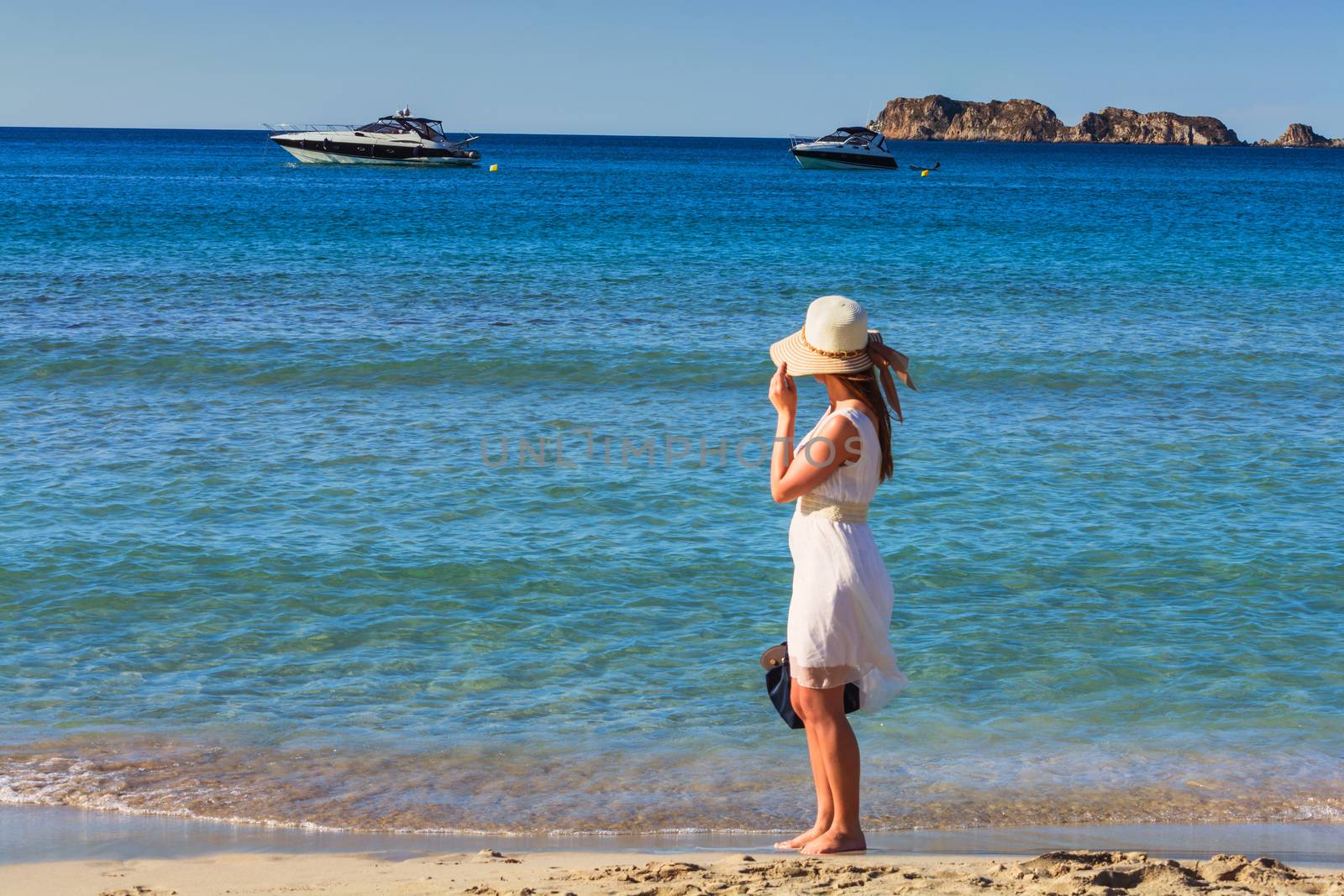 Young trendy woman relax on the beach. Happy island lifestyle. Holiday in paradise. In Hindergrund beach blue sky, crystal clear sea and two luxury motor yachts.