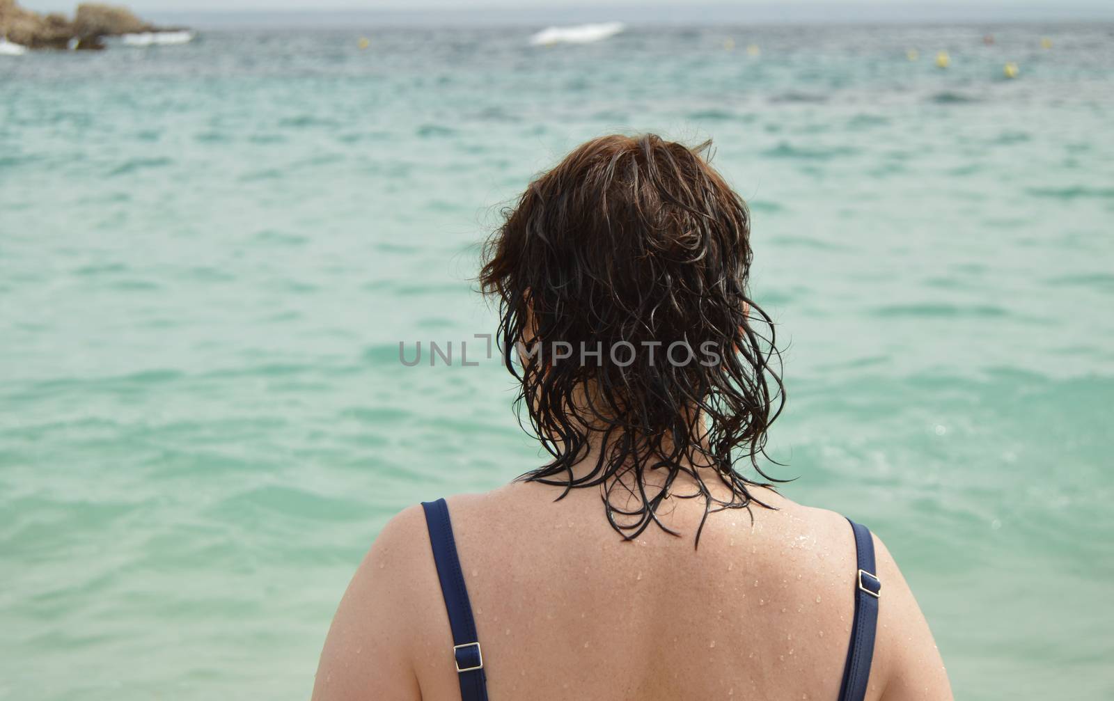 View of the back of an adult woman in a swimsuit with wet hair, standing on the beach and admiring the sun glare on the sea,