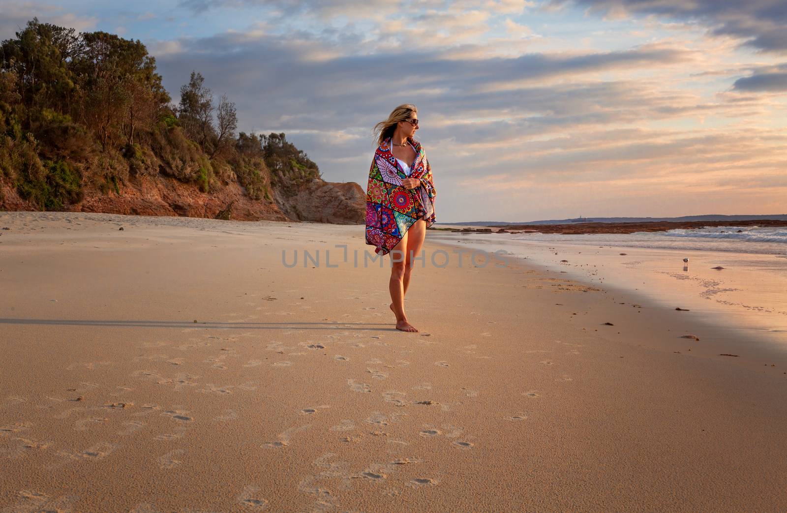 Female in bikini with a beach towel wrapped loosely around her walks along the beach in the early morning with soft breeze in her hair as she gazes towards the sun