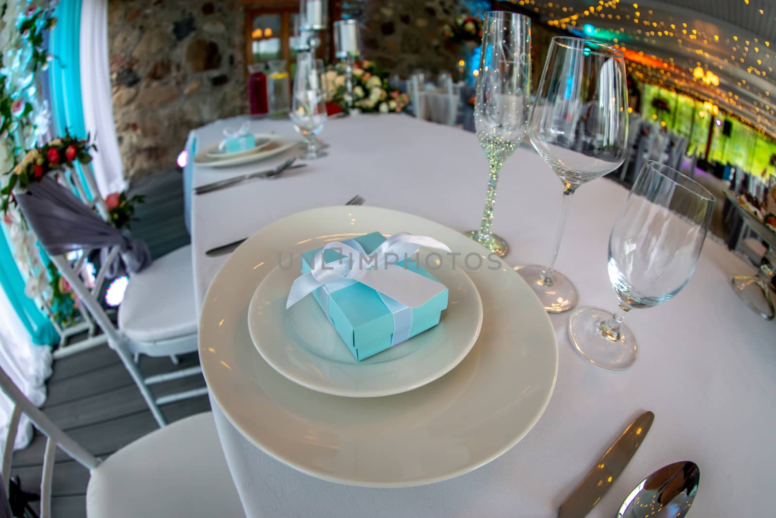 Table setting with handmade gift box on plate, glasses, forks, spoons and knives for wedding reception. Festive decorations and items for food, arranged on the table. Light blue gift box in plates. Shot with fisheye lens. 