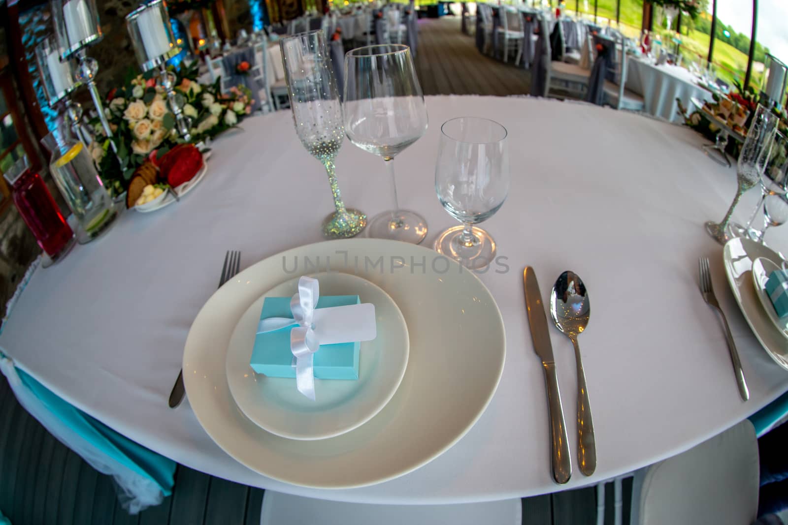 Table setting with handmade gift box on plate, glasses, forks, spoons and knives for wedding reception. Festive decorations and items for food, arranged on the table. Light blue gift box in plates. Shot with fisheye lens. 