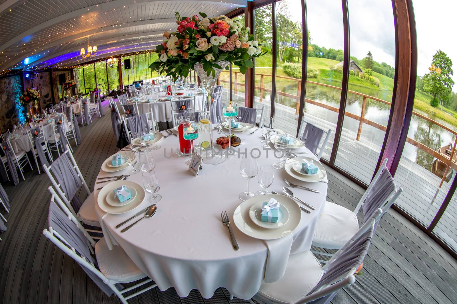 Table setting with handmade gift box on plate, bouquet of flowers, glasses, forks, spoons and knives for wedding reception. Guest tables in luxury decorated wedding banquet room. Shot with fisheye lens. 