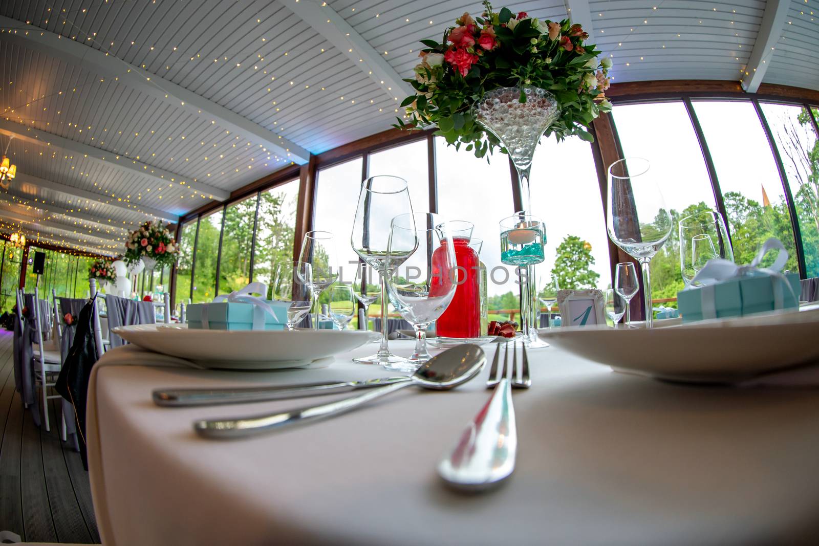 Table setting with handmade gift box on plate, bouquet of flowers, glasses, forks, spoons and knives for wedding reception. Guest tables in luxury decorated wedding banquet room. Shot with fisheye lens. 