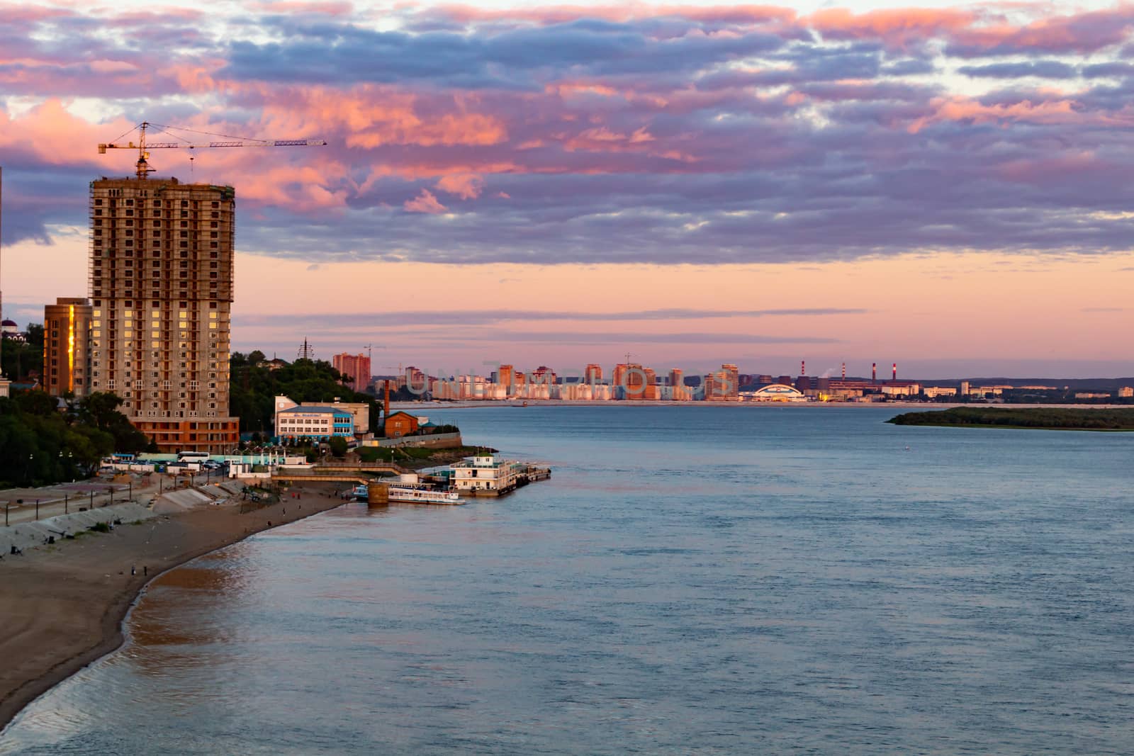 The sun sets over the Amur river. Buildings of the city of Khabarovsk in Golden pink. There are beautiful clouds in the sky.