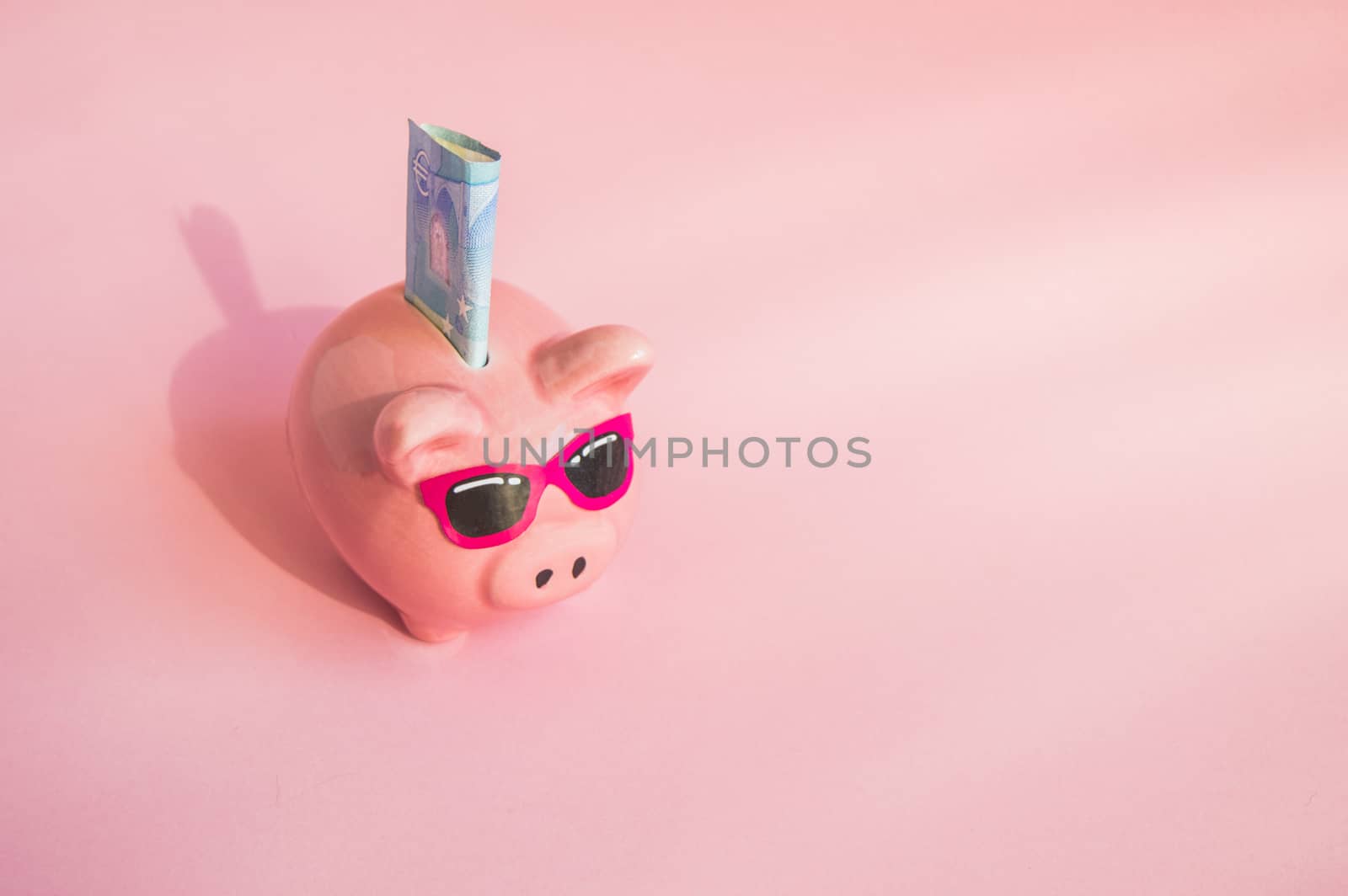Concept of saving money on your trip or vacation pig piggy Bank with sunglasses on the bill of euros on a pink background, place for text by claire_lucia